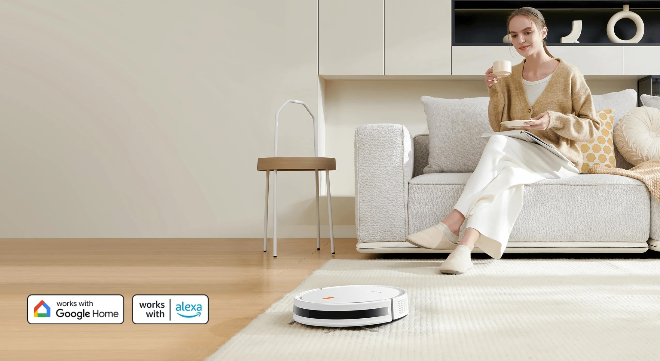Kobieta na kanapie trzymająca filiżankę. Robot odkurzacz na dywanie. Krzesło obok kanapy.