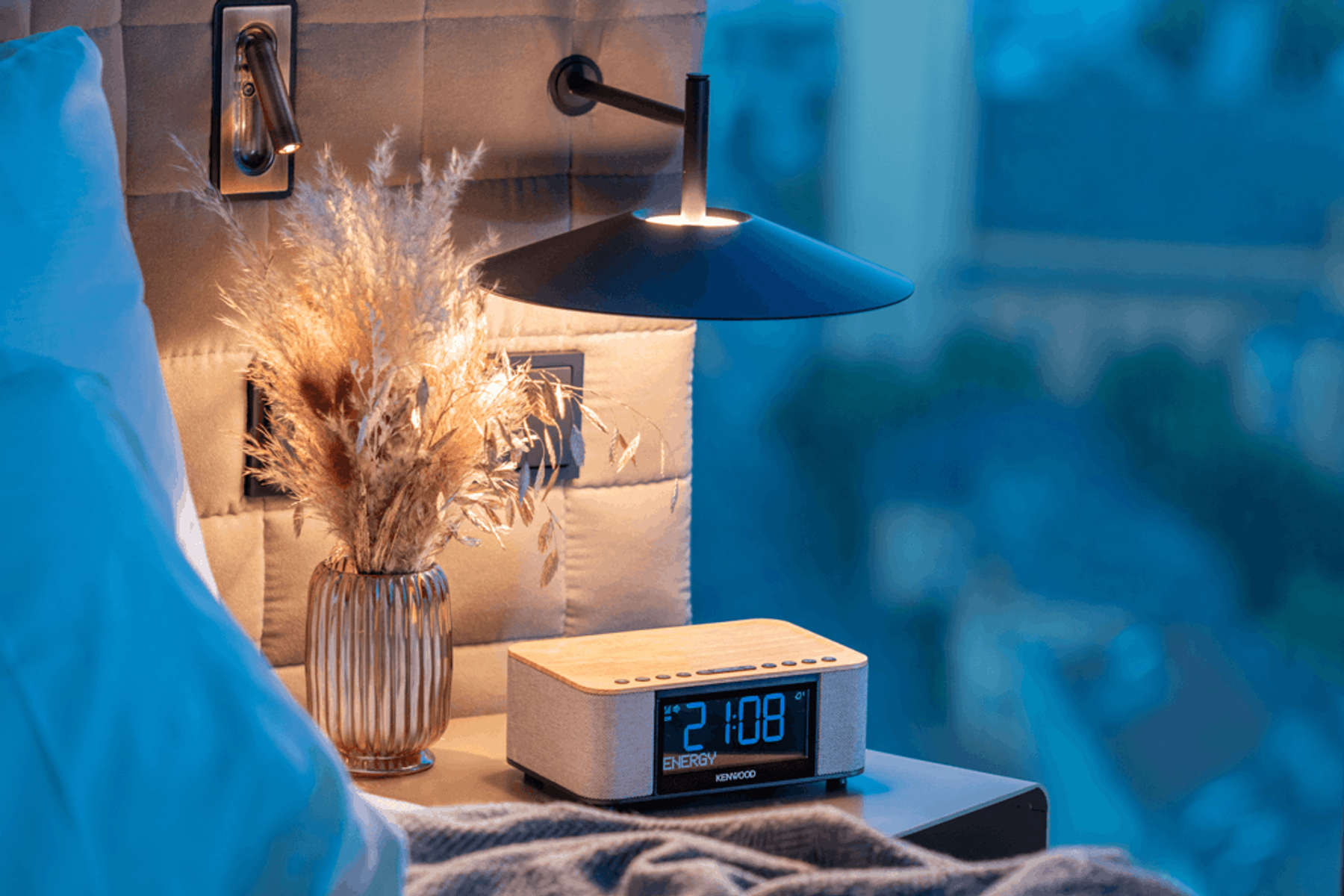 Scena przy łóżku z lampą, wazonem z roślinami i radiem z budzikiem. W tle rozmyty widok na miasto.