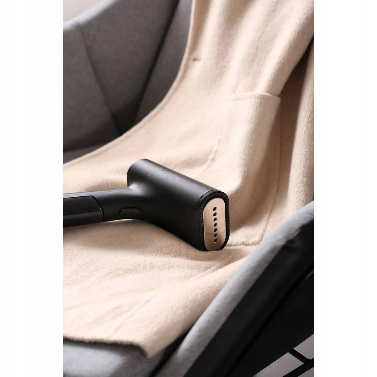 Un vaporizador de ropa negro está sobre un abrigo beige, que está sobre una silla gris. La cabeza del vaporizador tiene agujeros.