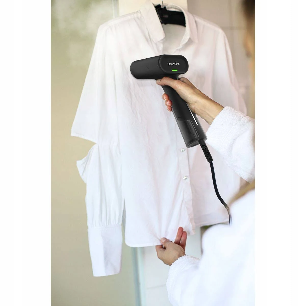 Una persona plancha una camisa blanca con un vaporizador negro. La camisa cuelga de un perchero. La persona lleva una bata blanca.