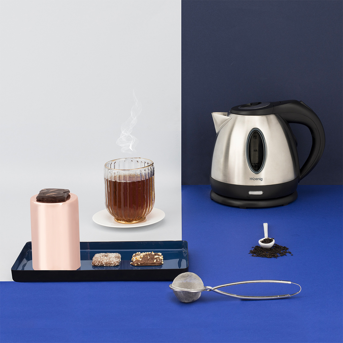 A tea setting with a glass of tea, a kettle, sweets, a tea infuser, and loose tea leaves on blue and white surfaces.