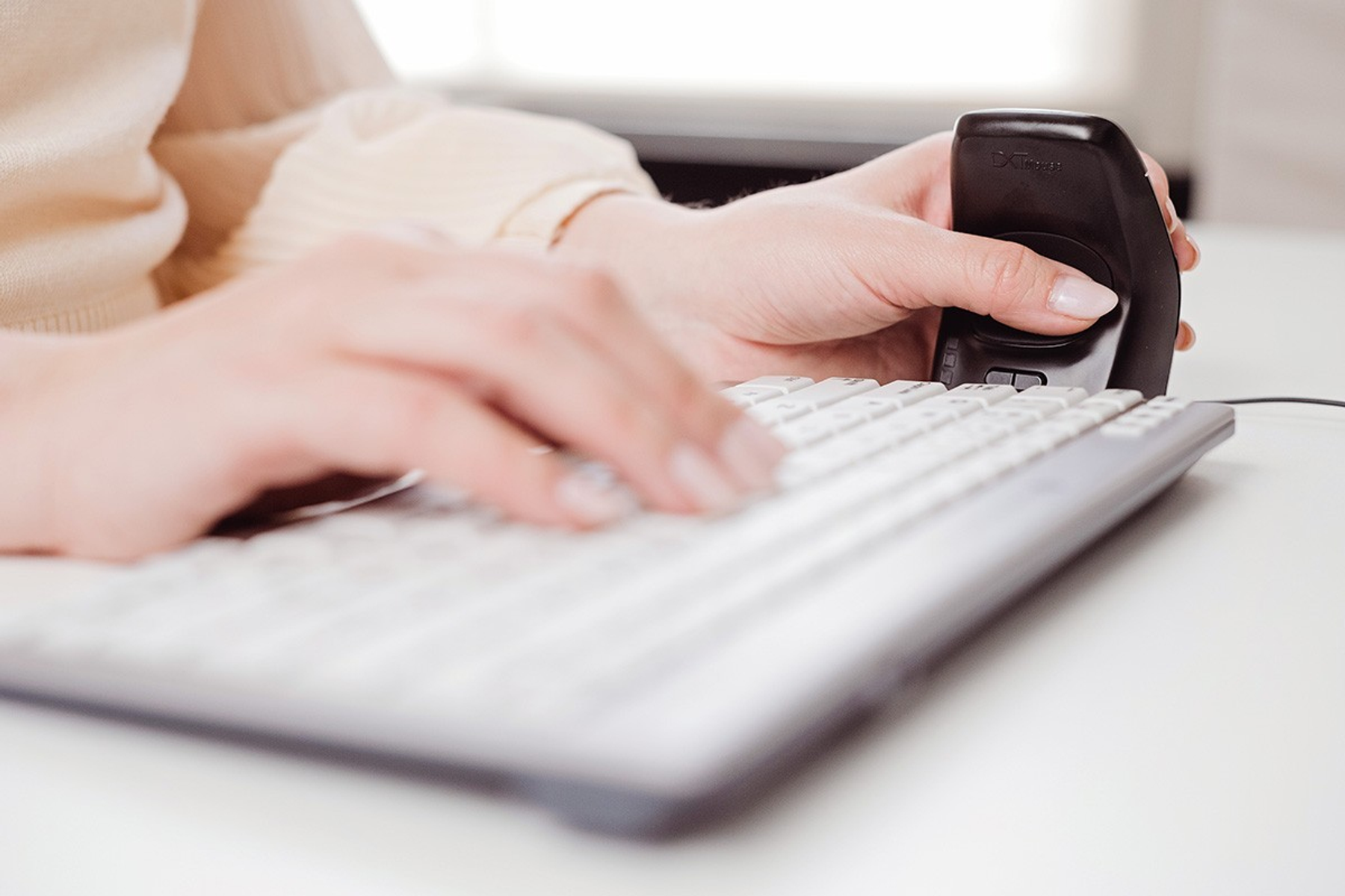 Person typing on white keyboard, holding a black trackball mouse. Hands and keyboard are in focus, desk is white.