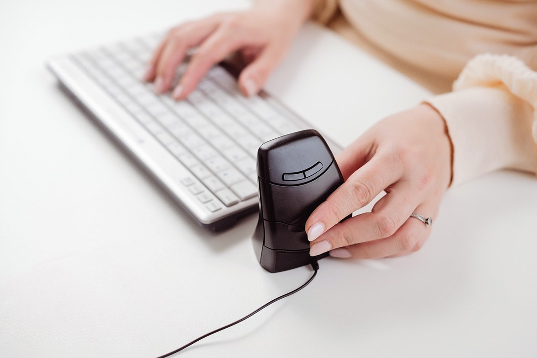 A person is using a computer. One hand is on the keyboard, the other holds a black vertical mouse. The background is white.