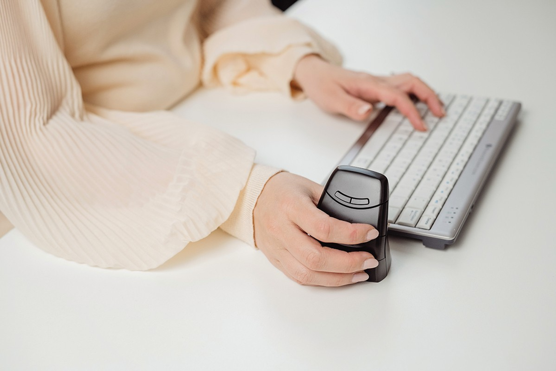 A person uses a vertical mouse and a keyboard. The person wears a cream-colored shirt. The desk is white.