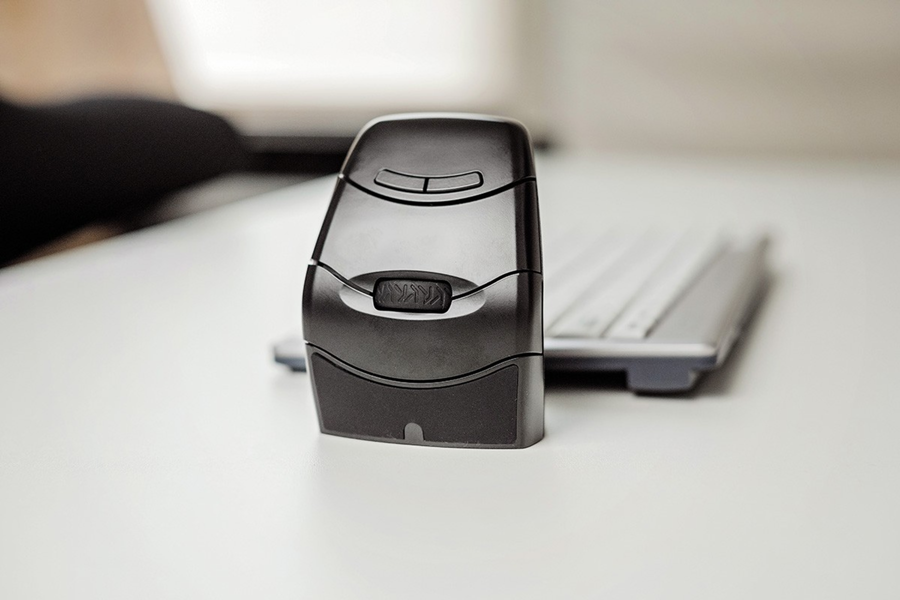 A black, ergonomic mouse stands upright on a white desk. A keyboard is in the background. The scene is well-lit and modern.