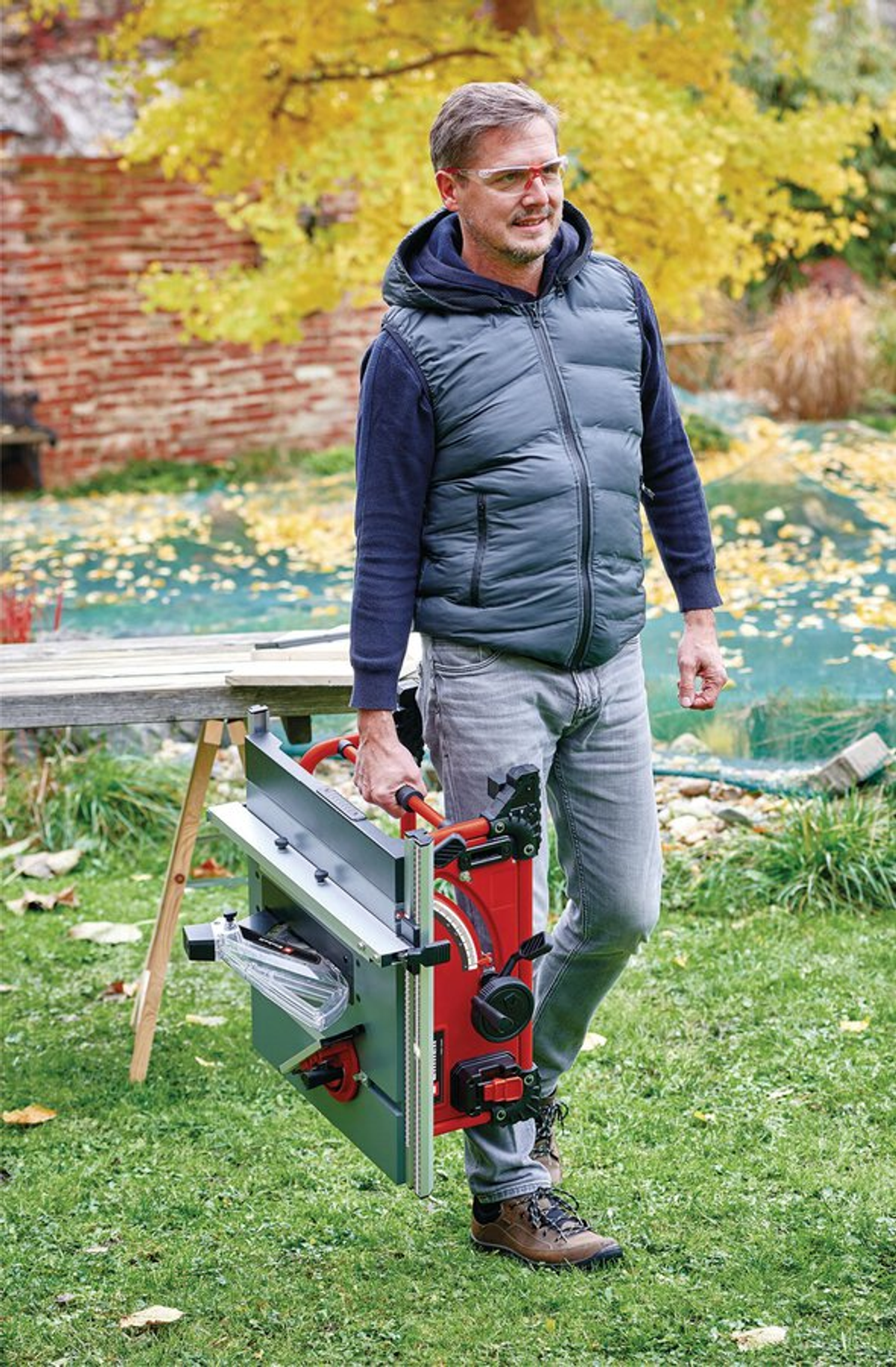 Ein Mann in einer Weste trägt eine rot-graue Tischkreissäge in einem Garten mit einem Holztisch, grünem Gras und einer Ziegelmauer.