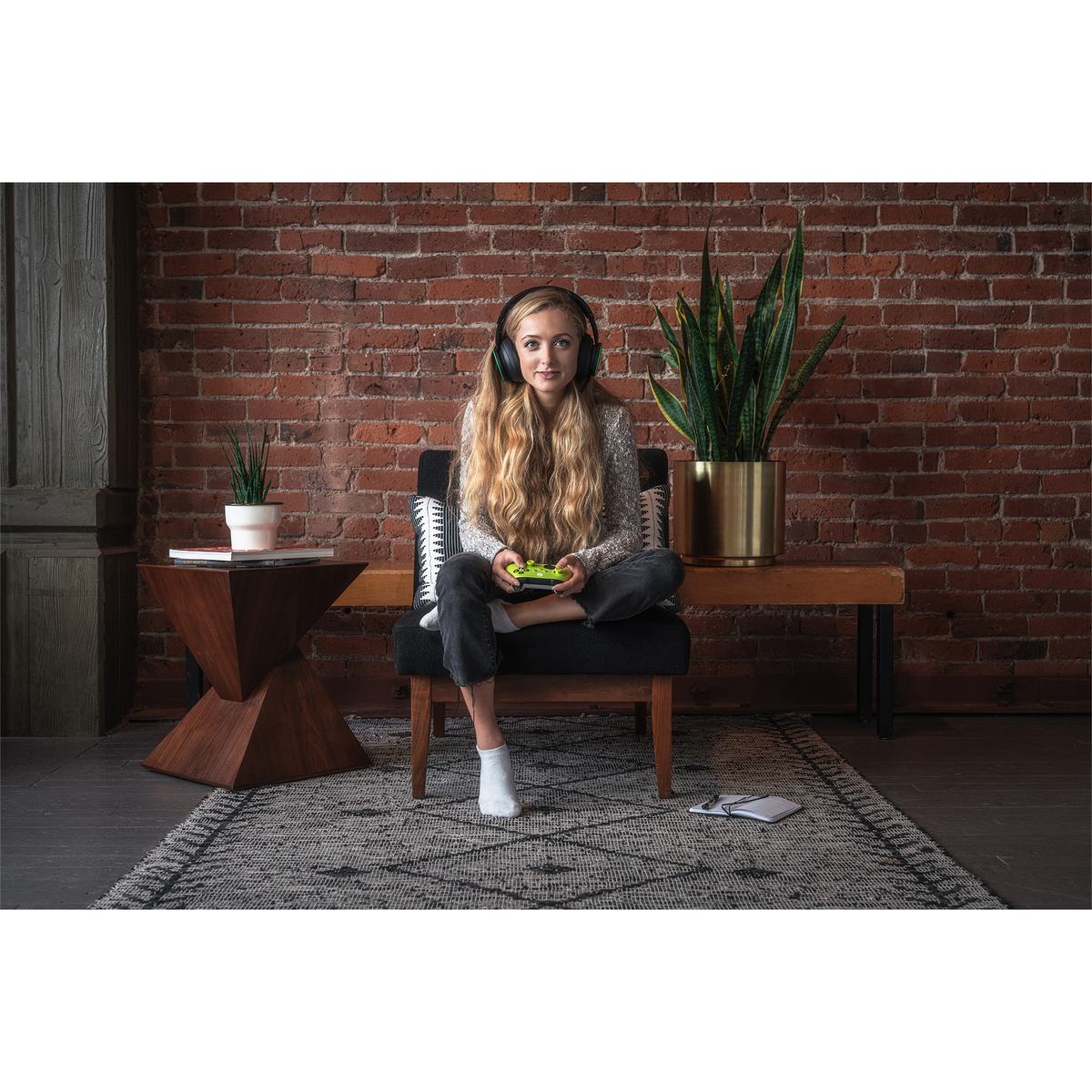 Eine junge Frau mit Kopfhörern sitzt in einem Stuhl und hält einen grünen Controller. Eine Backsteinmauer und Pflanzen sind im Hintergrund.