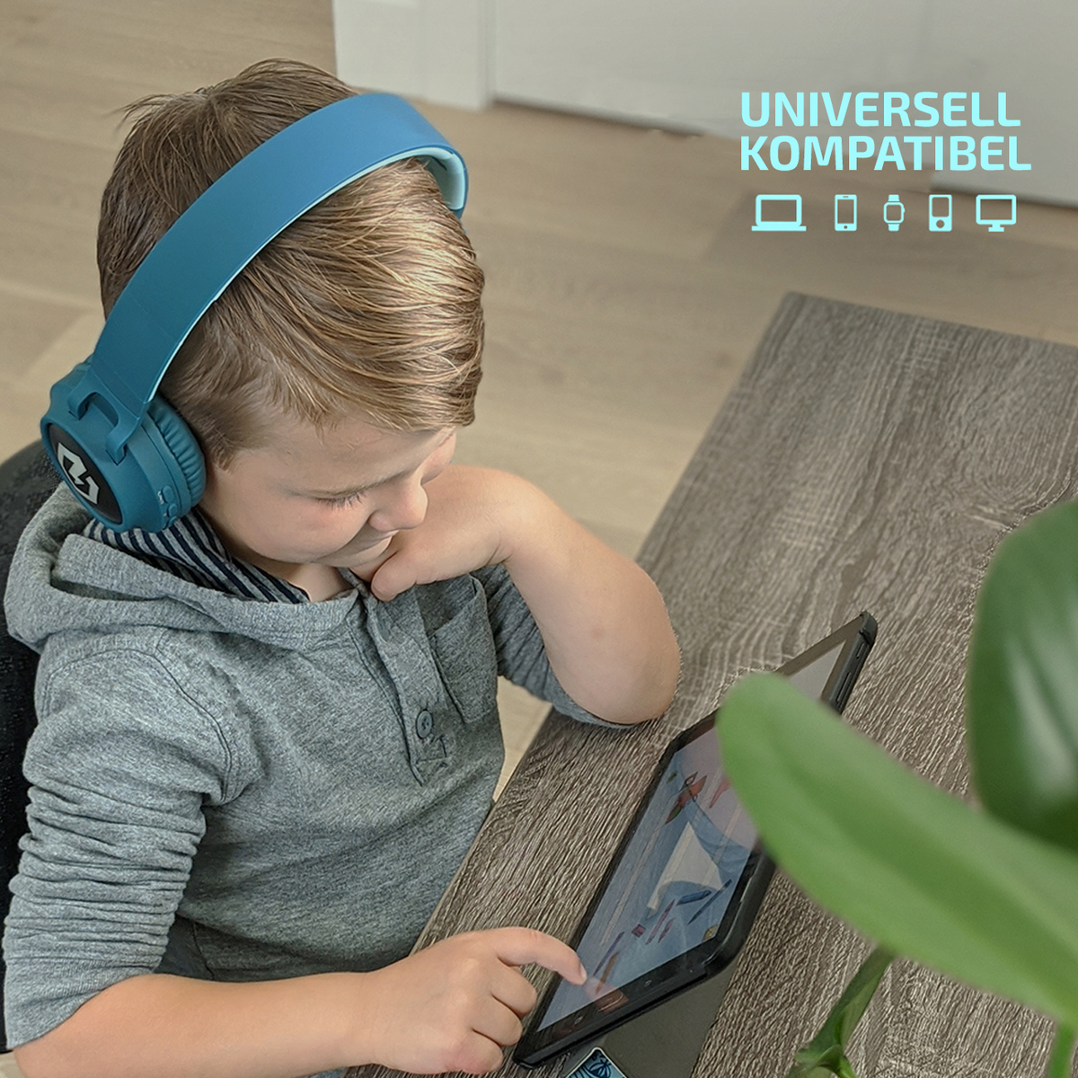 Un ragazzino con cuffie blu guarda un tablet. Sta toccando lo schermo. Tavolo in legno, pianta verde e testo sulla compatibilità.
