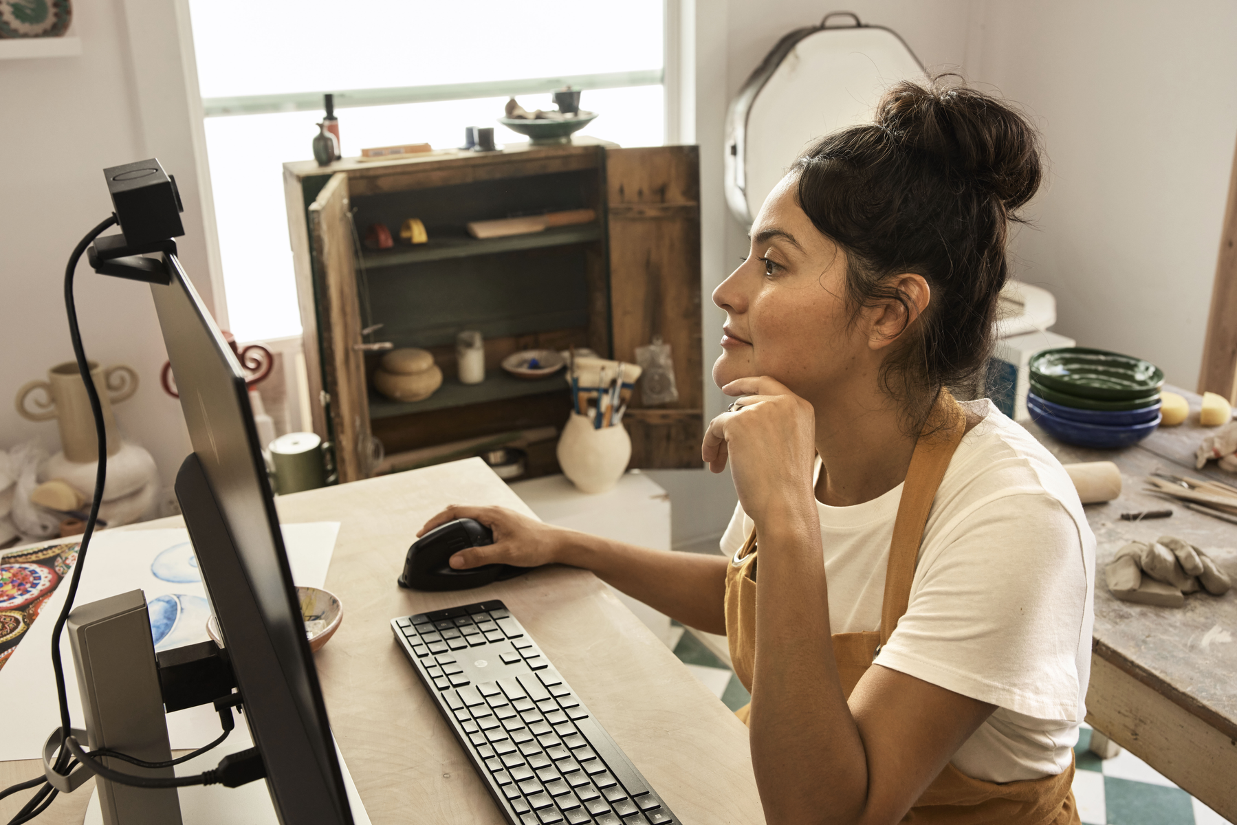 Kobieta siedzi przy biurku, pracując na komputerze. Ma na sobie białą koszulę i fartuch. Kamera internetowa znajduje się na monitorze.