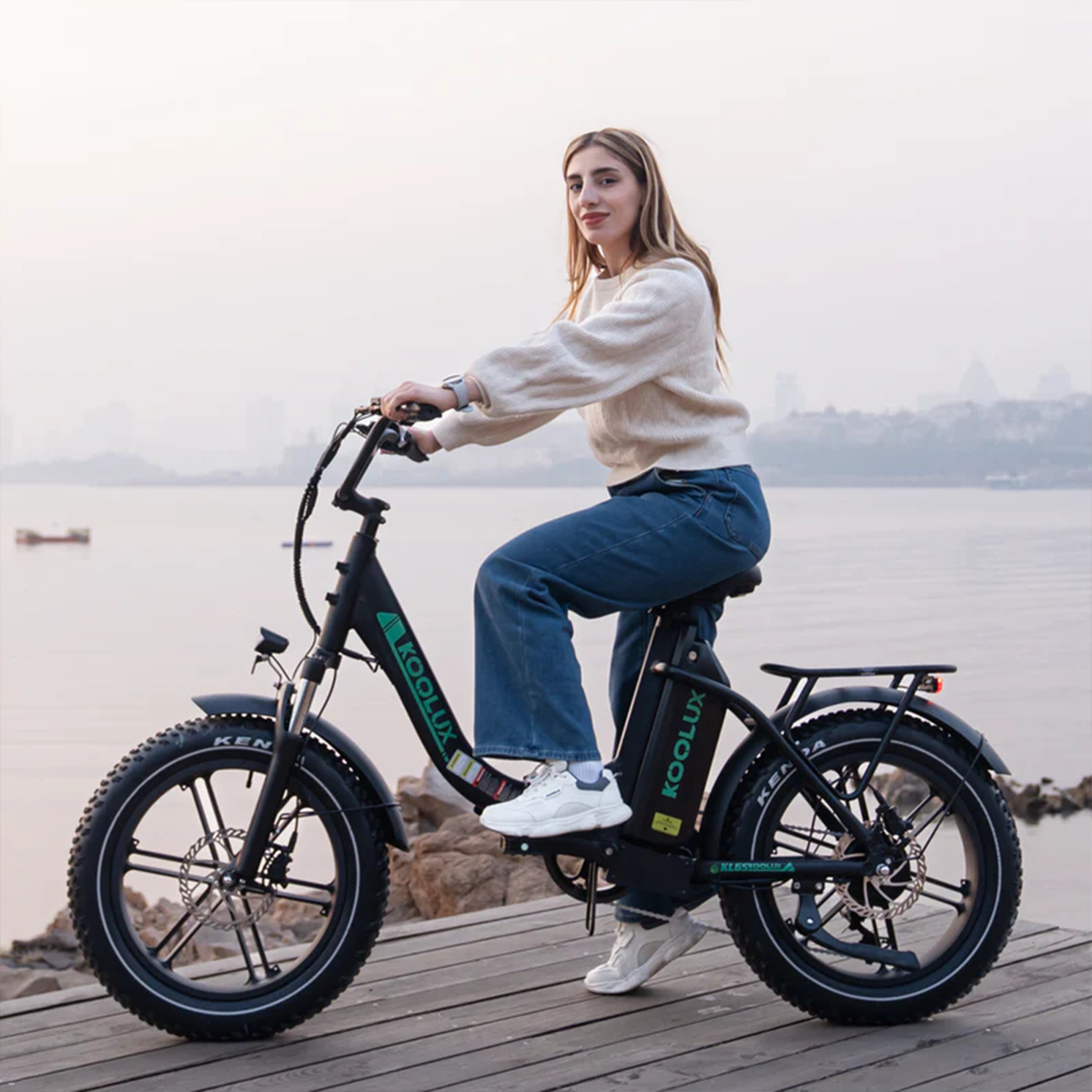 Kobieta siedzi na czarnym rowerze elektrycznym. Ma na sobie biały sweter i niebieskie dżinsy. W tle woda i miasto.