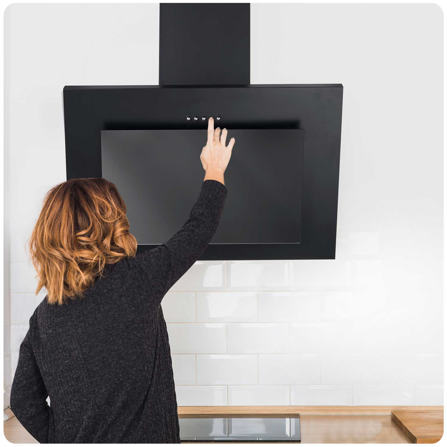 Ręka kobiety sięga po czarny okap. Ma na sobie ciemny sweter i rude włosy. Biała kafelkowa ściana w tle.