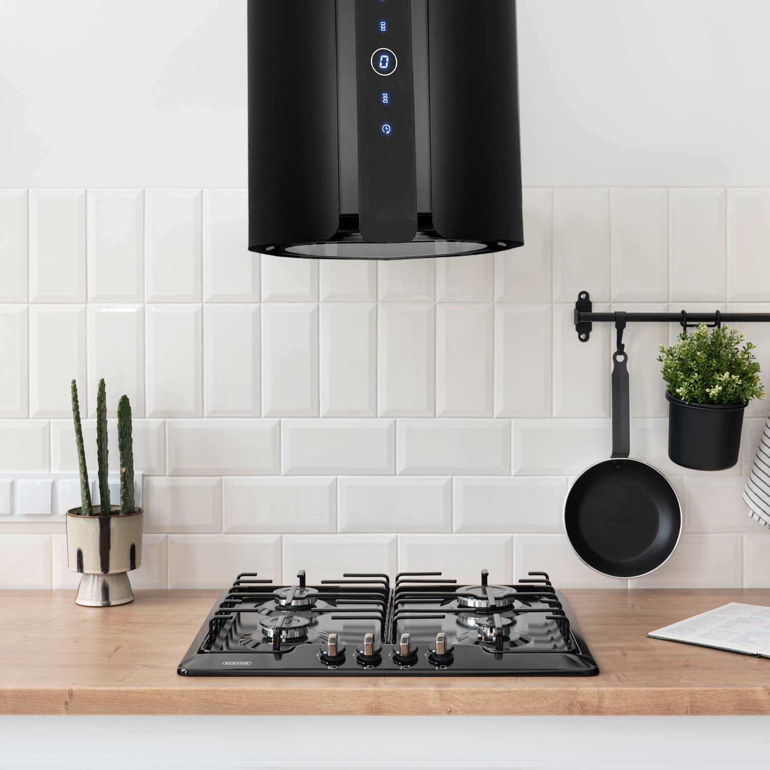 Scena kuchenna z czarnym okapem, płytą grzejną, patelnią i rośliną doniczkową. Biała ściana z płytek i drewniany blat. Kaktus po lewej.