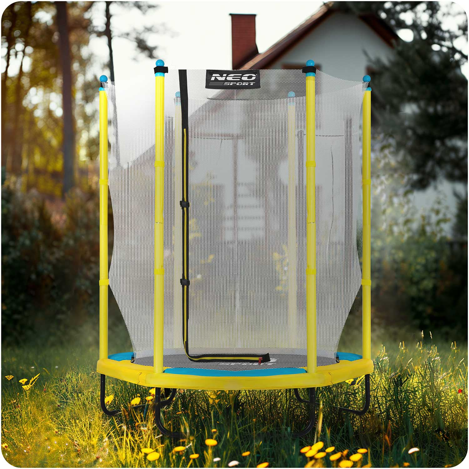 Żółta trampolina z siatką bezpieczeństwa stoi na trawie, w tle dom. Promienie słoneczne przedzierają się przez drzewa.