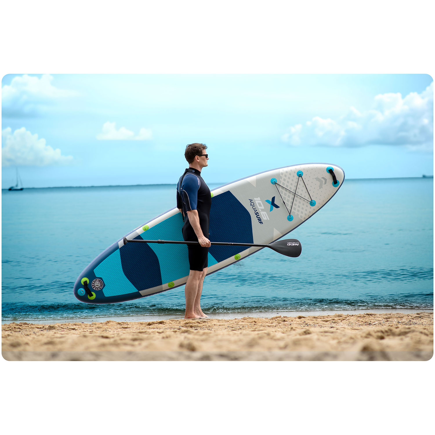 Mężczyzna w okularach przeciwsłonecznych trzyma deskę do wiosłowania na plaży, z morzem i niebem w tle. Deska ma markę Aquasurf.