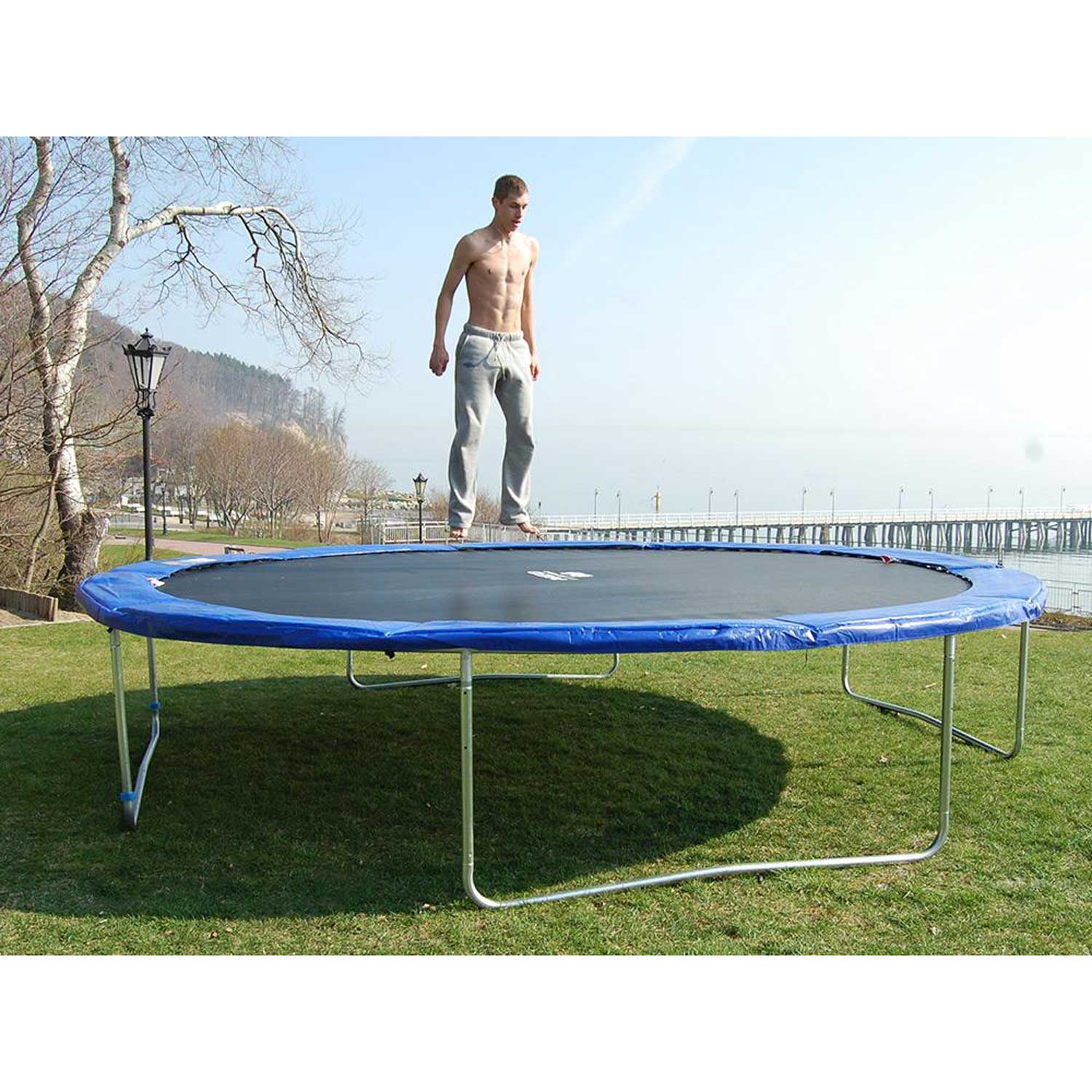 Mężczyzna skacze na trampolinie. Jest na trawie blisko wody. W tle molo i drzewa. Krawędź jest niebieska.