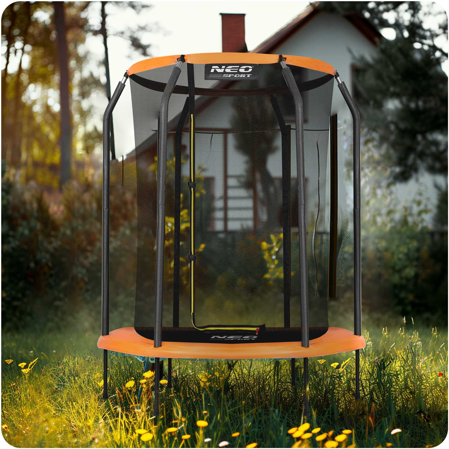 Trampolina z pomarańczową ramą i czarną siatką stoi na polu trawy. W tle dom i drzewa.