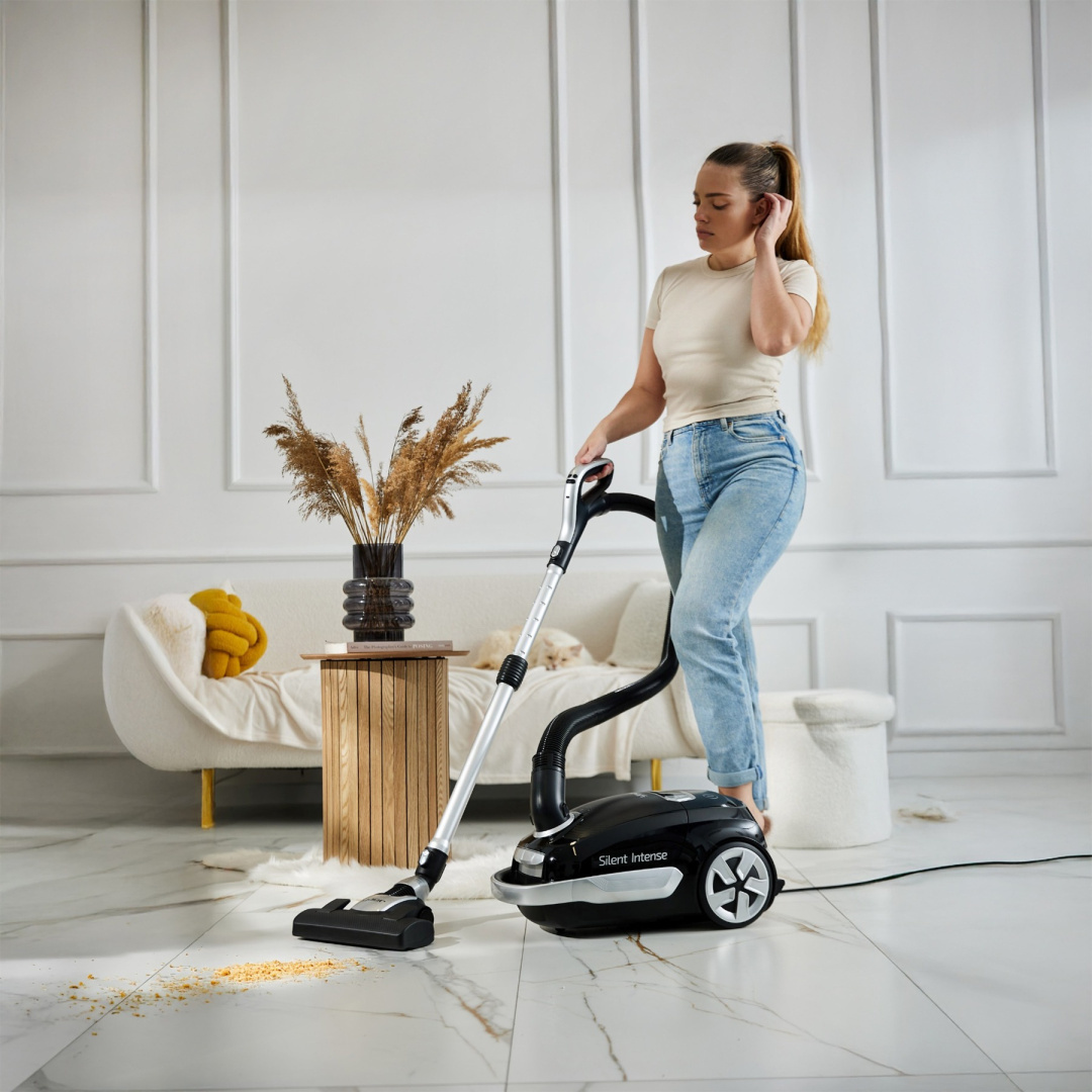 Kobieta odkurza białą, marmurową podłogę. Czarny odkurzacz jest w użyciu. W tle sofa, kot i dekoracje.