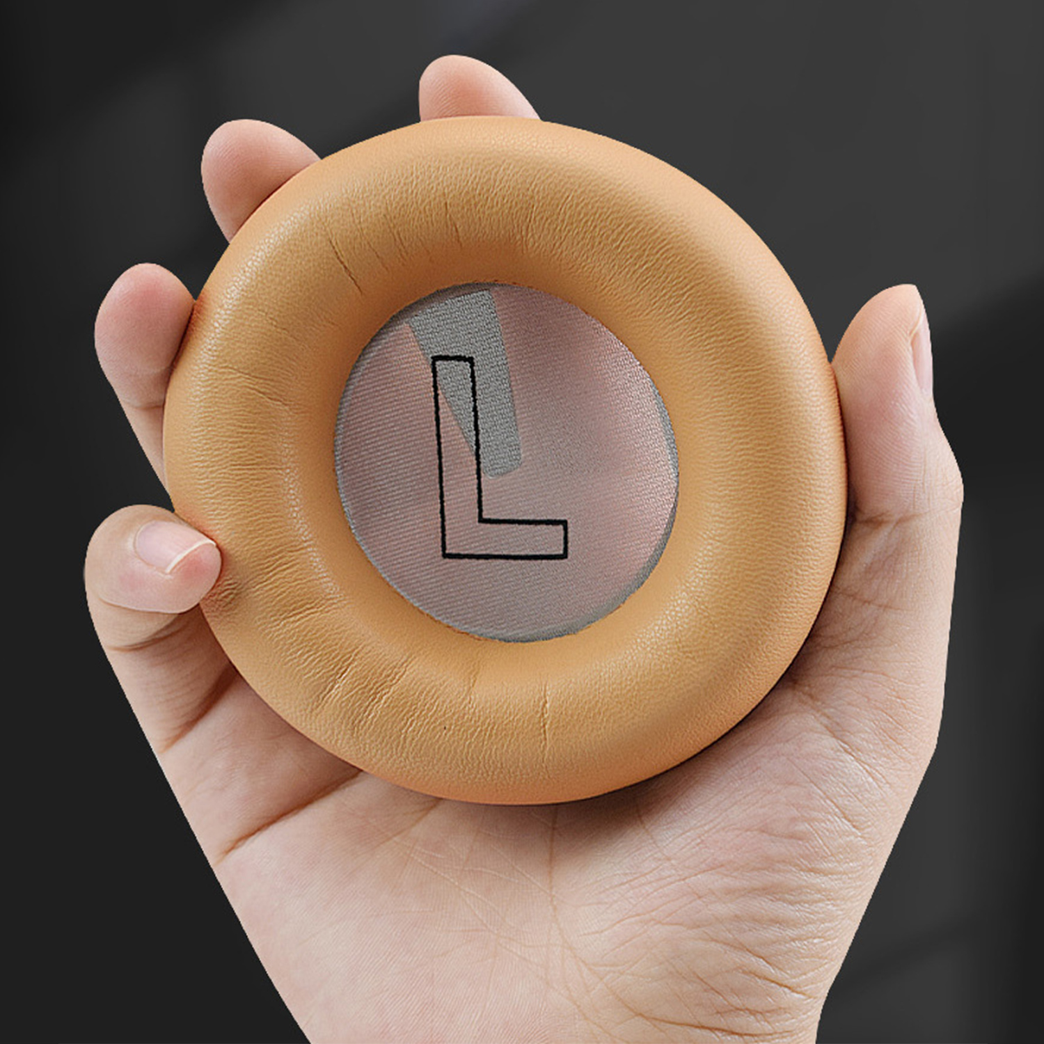 Dłoń trzyma beżową słuchawkę. Środek ma siatkę z czarną literą 'L'. Tło jest ciemne.