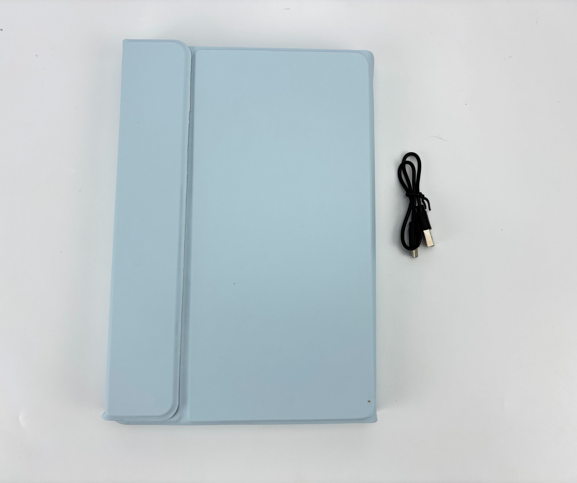 Eine hellblaue Tablet-Hülle und ein schwarzes Ladekabel liegen auf einer weißen Oberfläche. Die Hülle ist geschlossen.