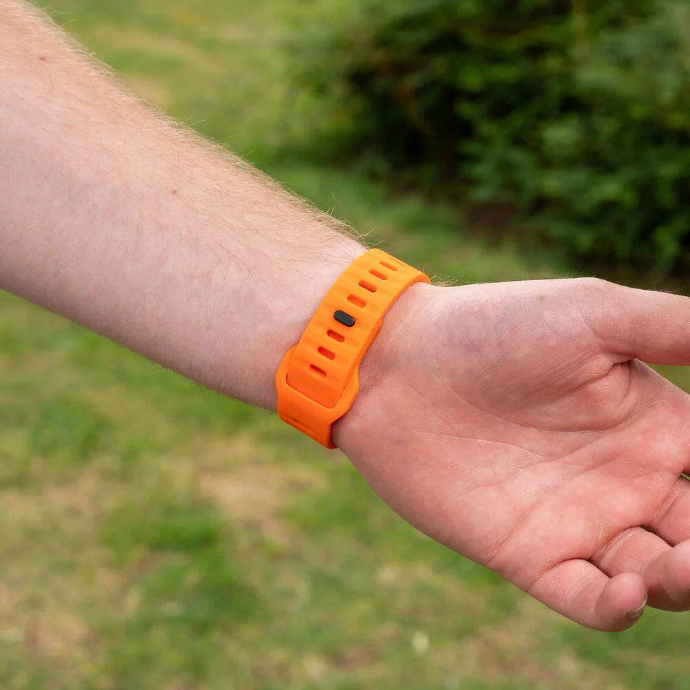 Ein Handgelenk mit einem orangefarbenen Smartband. Die Hand ruht, mit grünem Hintergrund aus Gras und Büschen.
