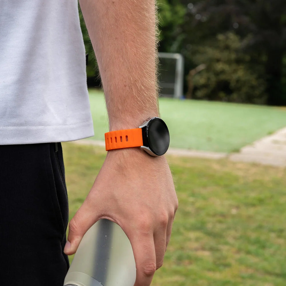 Eine Person trägt eine Smartwatch mit orangefarbenem Armband. Die Uhr befindet sich am Handgelenk. Der Hintergrund ist ein grünes Feld.