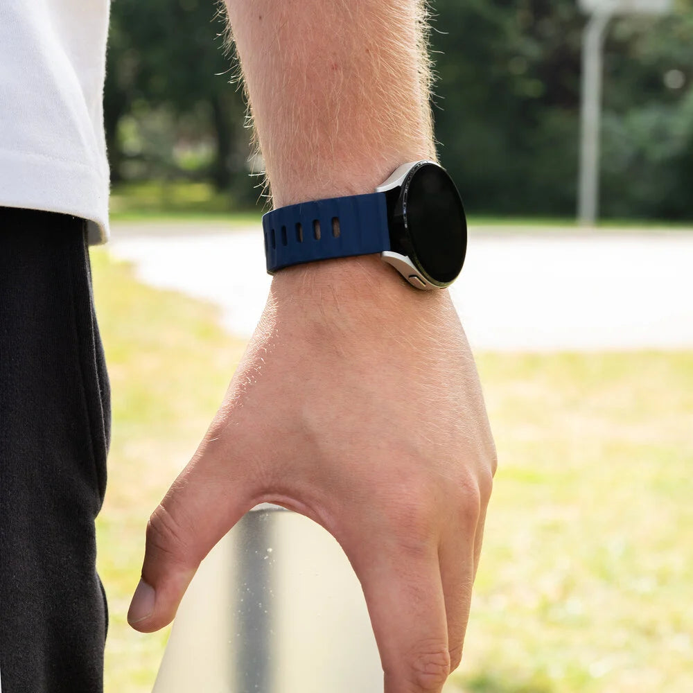 Eine Person, die eine blaue Smartwatch trägt. Die Uhr befindet sich am Handgelenk. Der Hintergrund zeigt einen Basketballplatz.