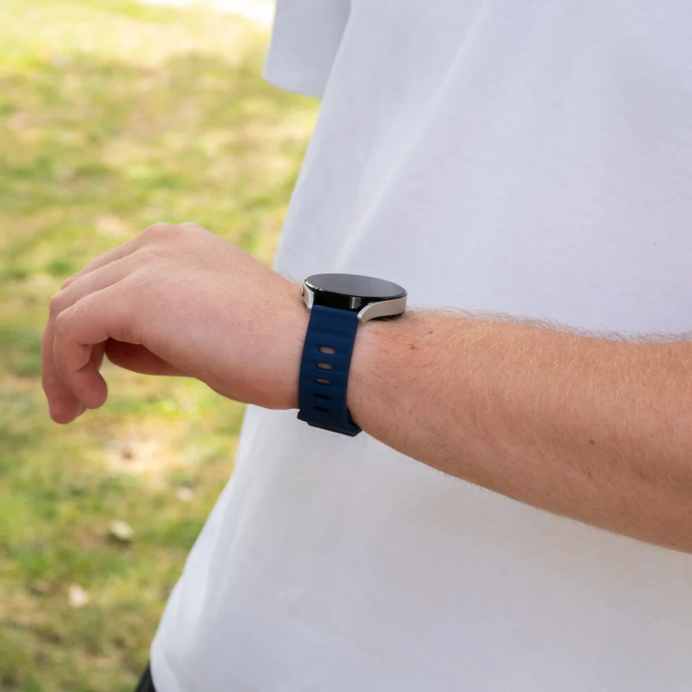Eine Person trägt eine Smartwatch mit blauem Armband. Die Uhr befindet sich am linken Handgelenk. Der Hintergrund ist verschwommen grün.