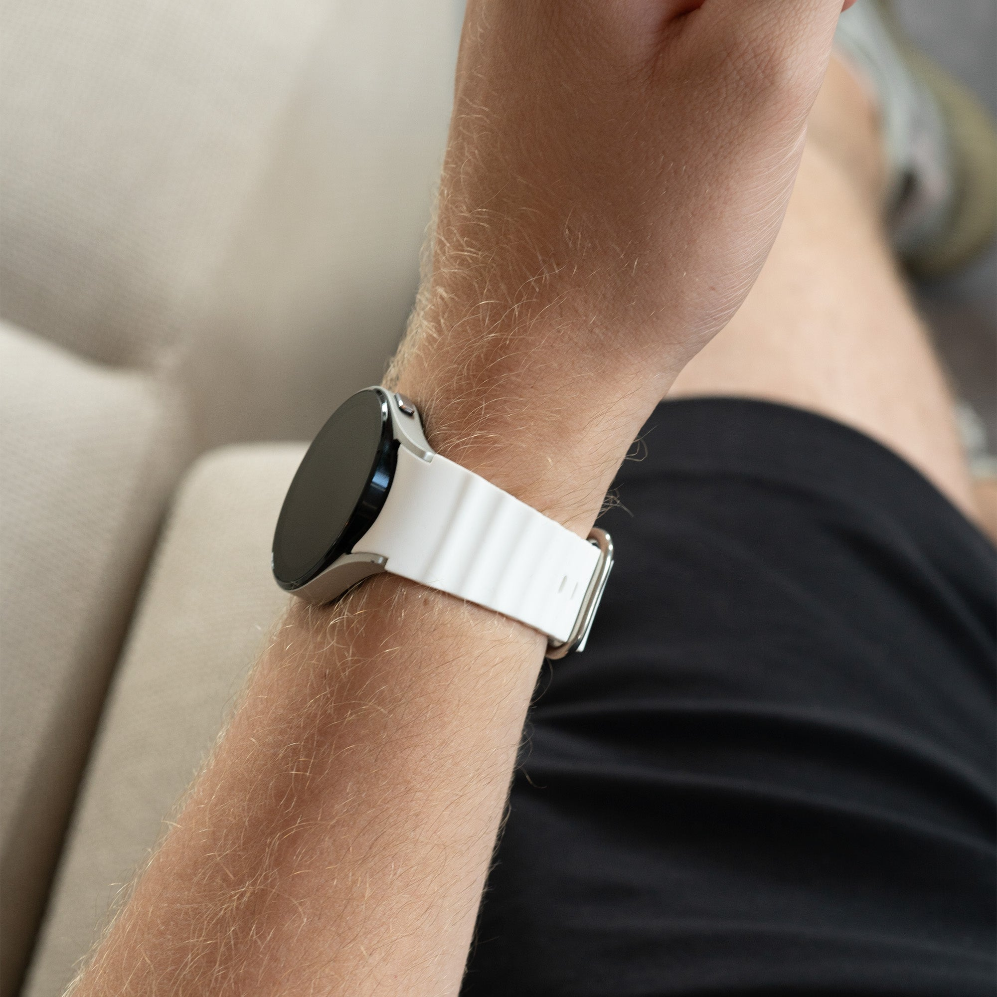 Ein Arm mit einer weißen Smartwatch mit schwarzem Bildschirm. Der Arm ruht. Schwarze Shorts und ein helles Sofa sind im Hintergrund.