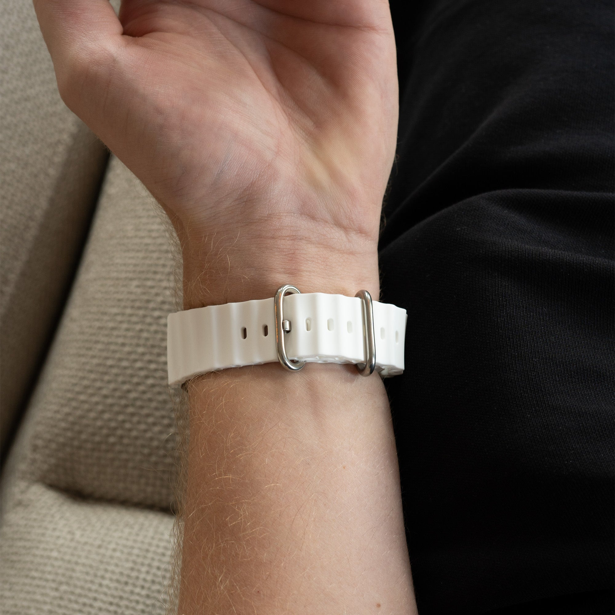 Ein Handgelenk mit einem weißen Armband ist zu sehen. Das Armband hat ein wellenförmiges Design und silberne Verschlüsse. Die Person trägt ein schwarzes Hemd.
