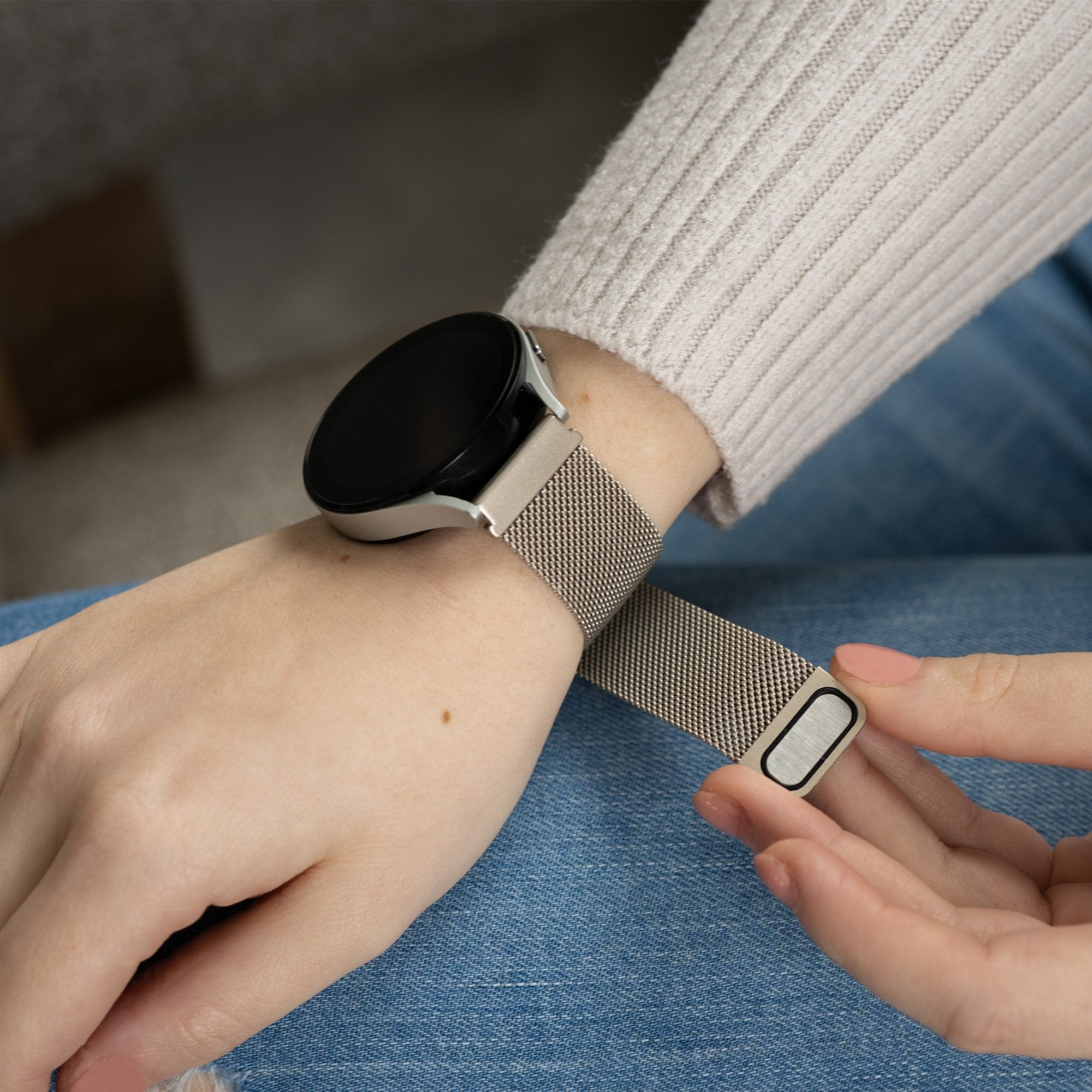 Eine Person befestigt eine silberne Smartwatch mit einem Mesh-Armband an ihrem Handgelenk. Sie trägt einen beigen Pullover und blaue Jeans.