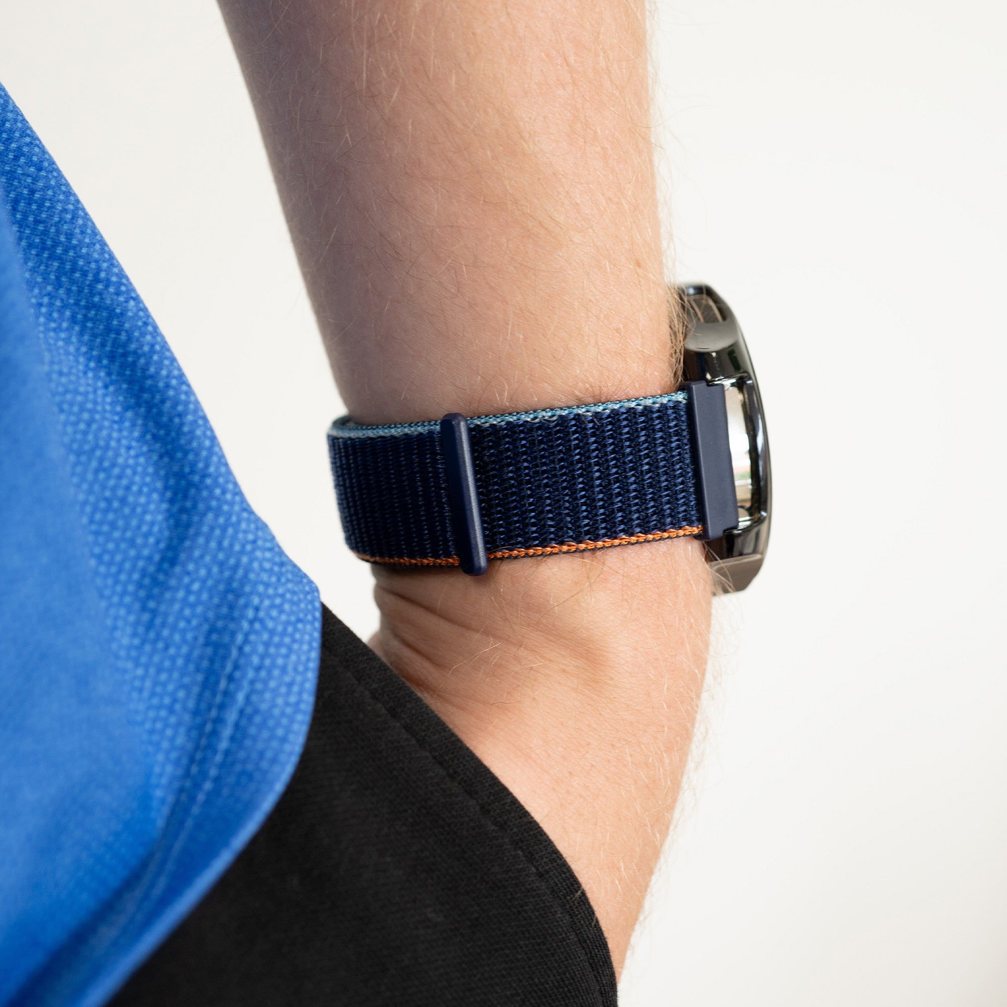 Der Arm einer Person mit einem blauen Armband und einer schwarzen Uhr. Ein blaues Hemd ist sichtbar.
