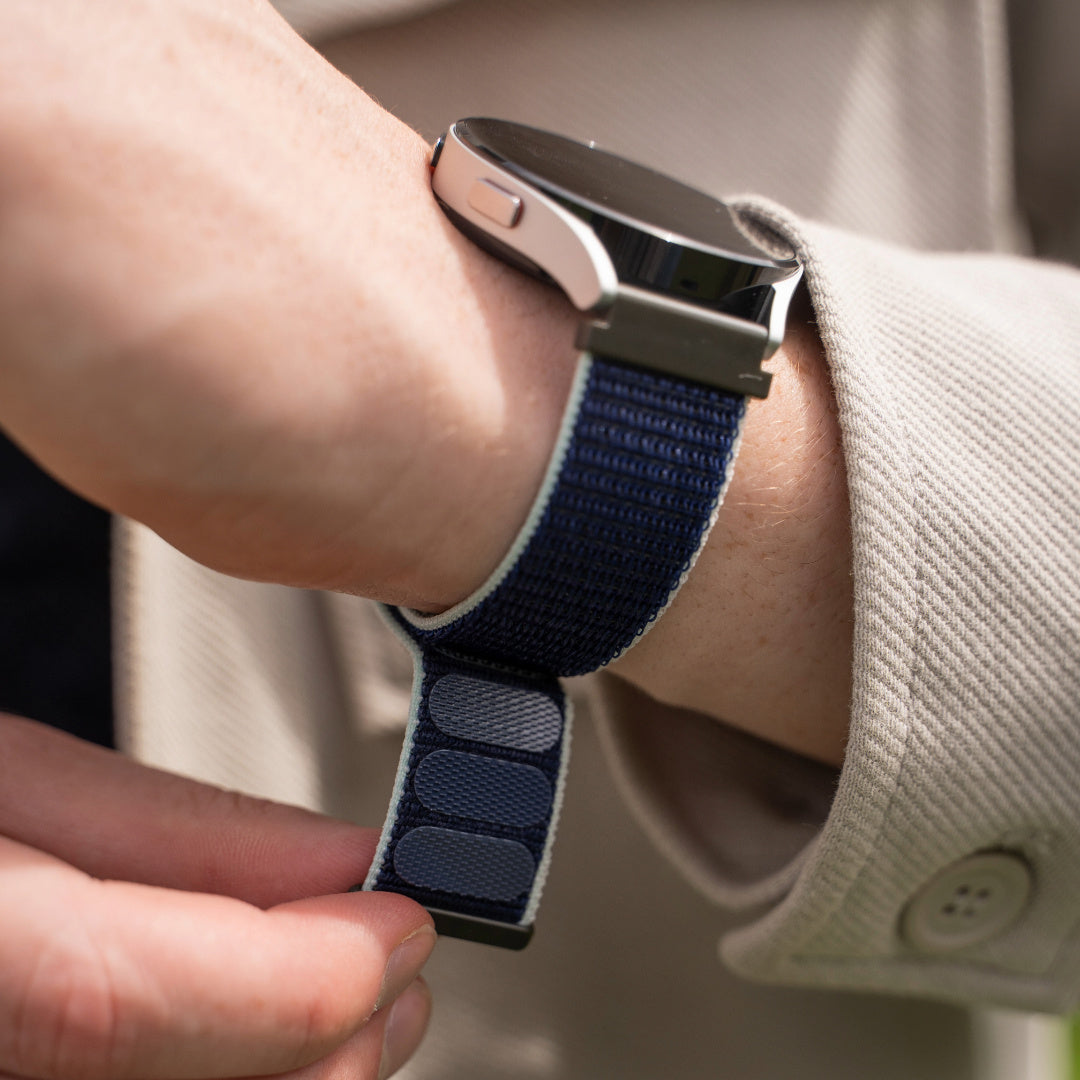 Ein Handgelenk mit einer Smartwatch. Die Uhr hat ein marineblaues Armband. Die Person trägt eine beige Jacke.