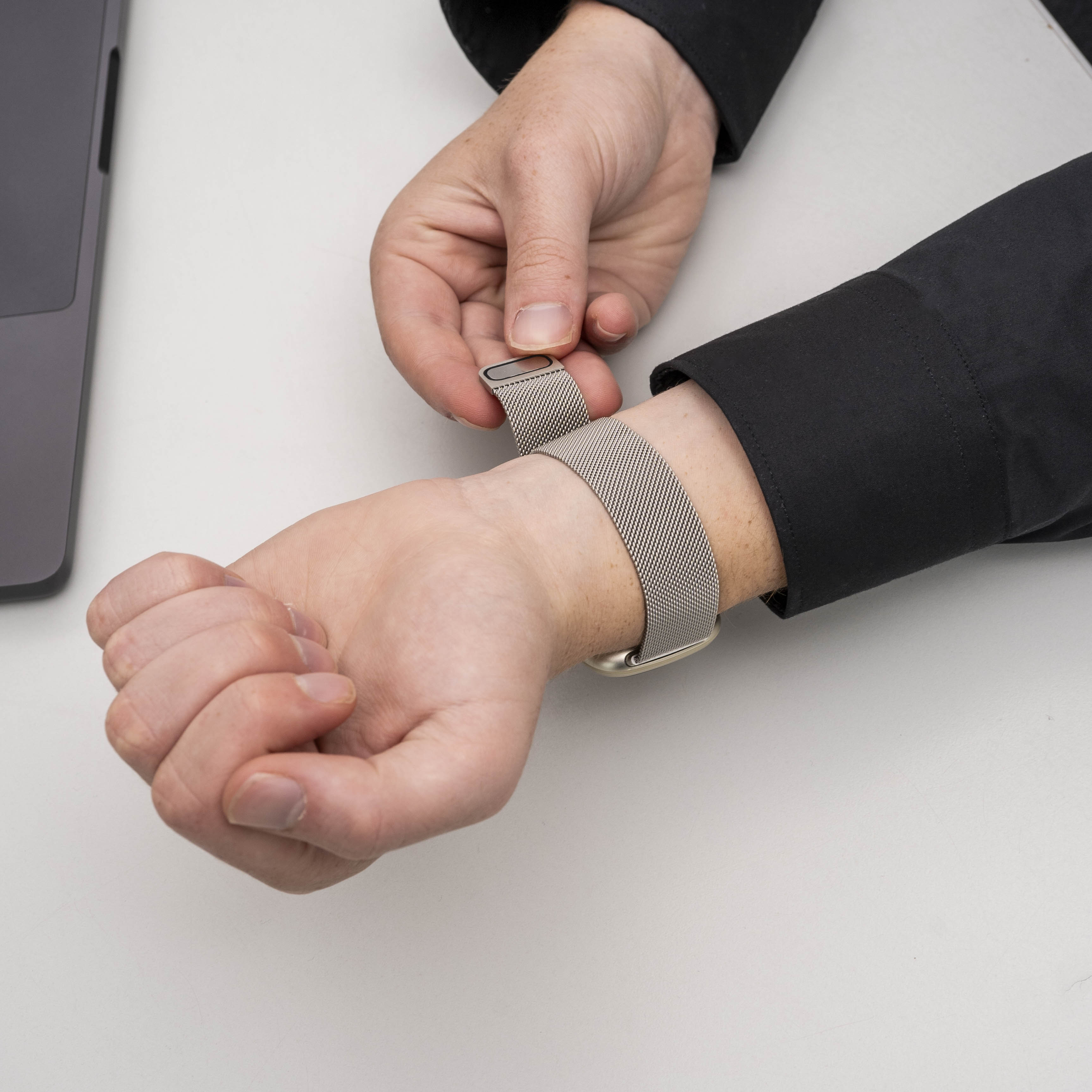 Die Hand einer Person befestigt ein metallisches Mesh-Uhrenarmband am Handgelenk. Links ist ein Laptop zu sehen.