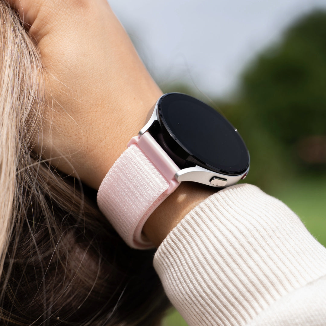 Handgelenk mit silberner Smartwatch, hellrosa Armband und weißem Pulloverärmel. Verschwommener grüner Hintergrund.