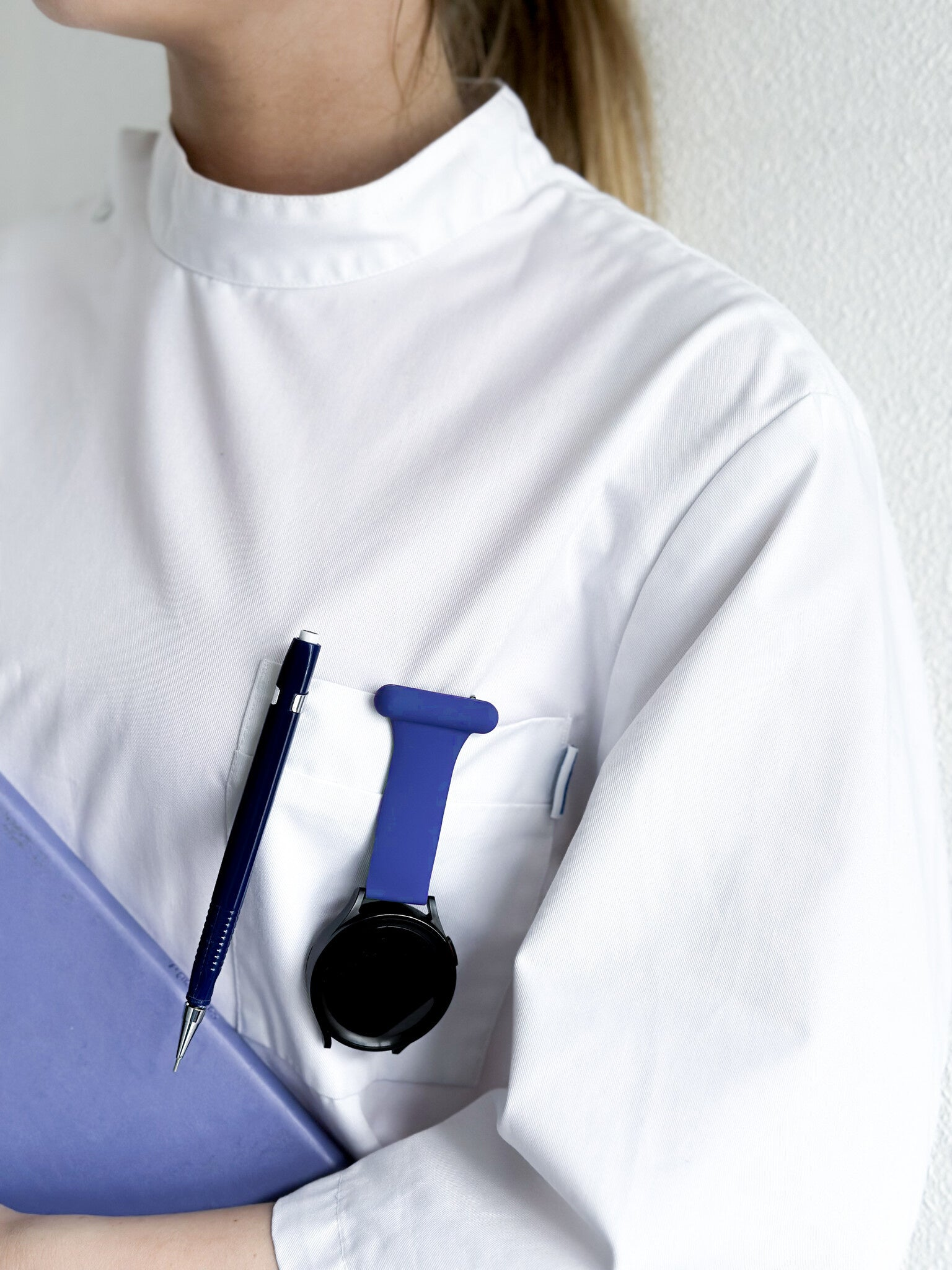 Eine Person in einem weißen Kittel hält einen lila Ordner. Ein Stift und eine blaue Uhr sind in der Tasche. Der Hintergrund ist eine weiße Wand.