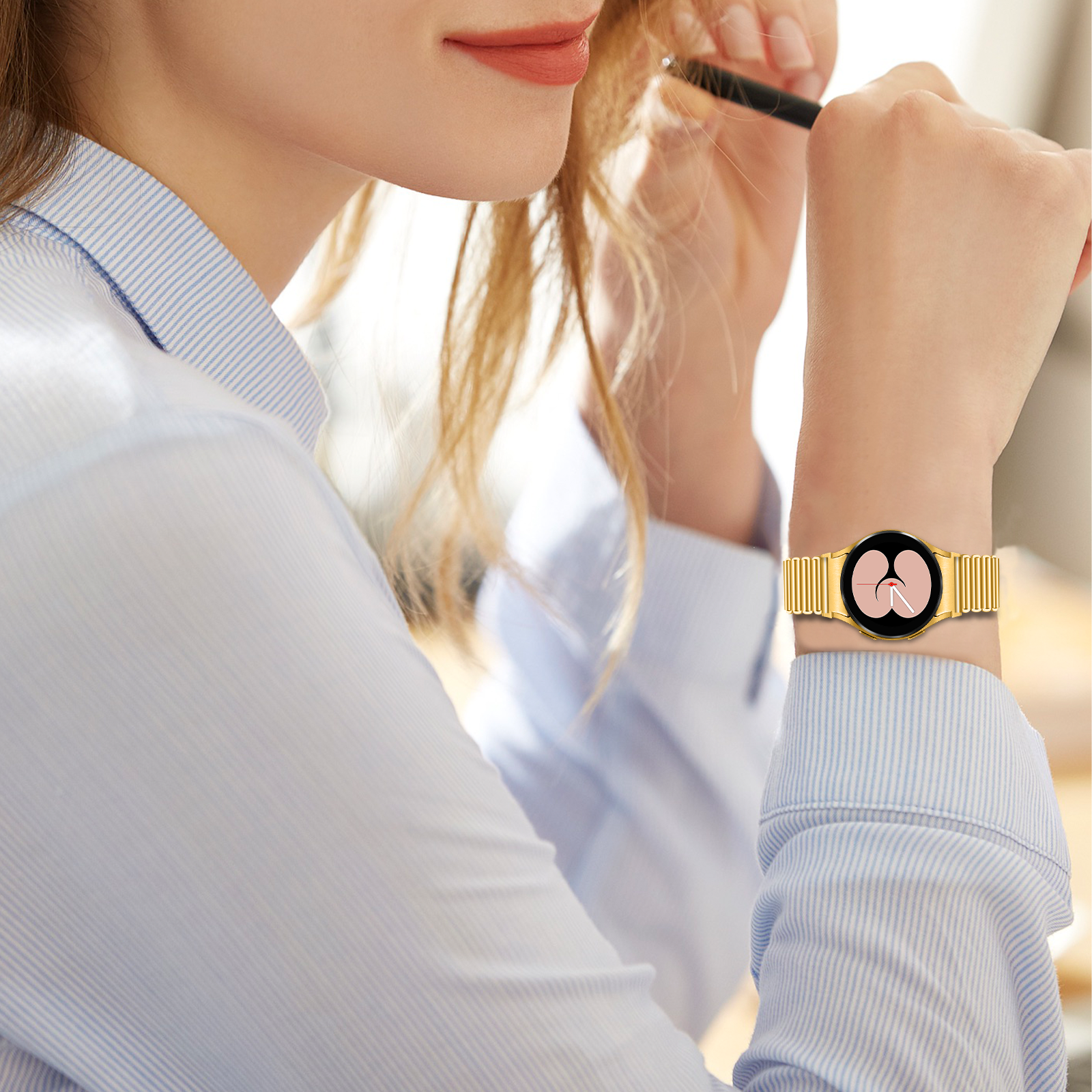 Eine Frau mit einer goldenen Uhr mit schwarzem Zifferblatt. Sie hält einen Stift in der Nähe ihrer Haare und trägt ein blau-weiß gestreiftes Hemd.