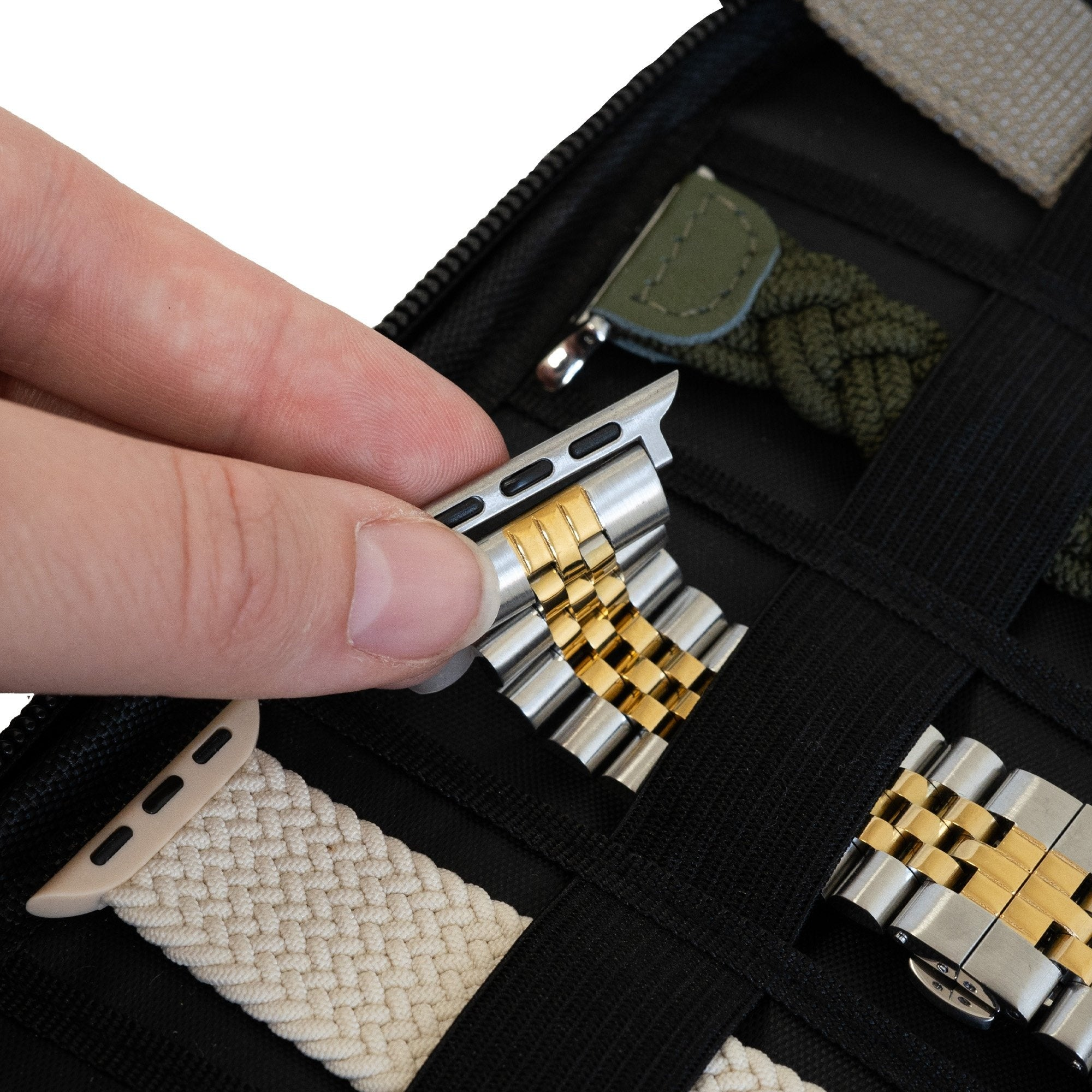 Eine Hand hält ein silbernes und goldenes Uhrenarmband über einem schwarzen Etui. Andere Armbänder und Zubehörteile sind im Hintergrund.