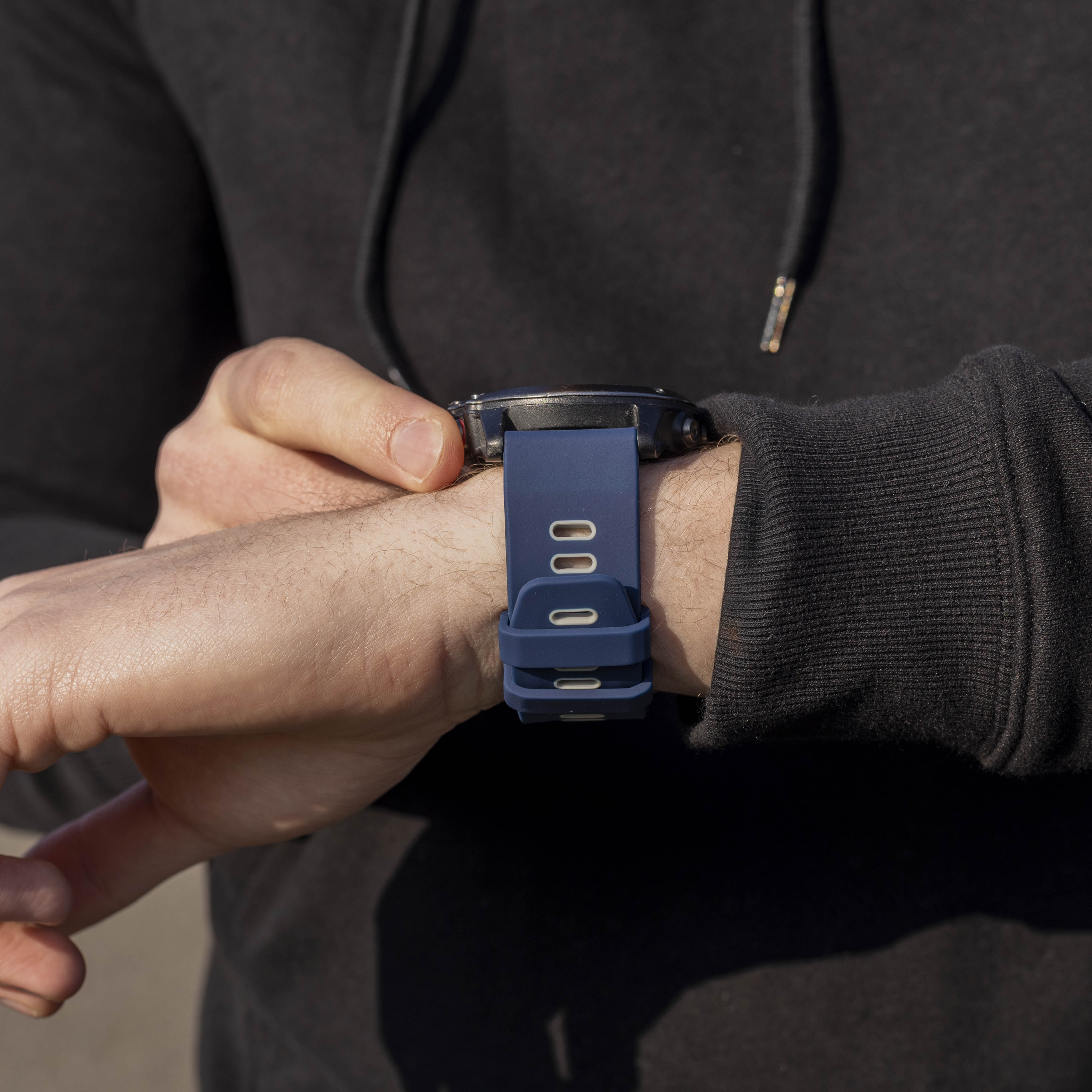 Eine Person in einem dunklen Kapuzenpullover betrachtet eine marineblaue Uhr an ihrem Handgelenk. Die Uhr hat ein blaues Armband.