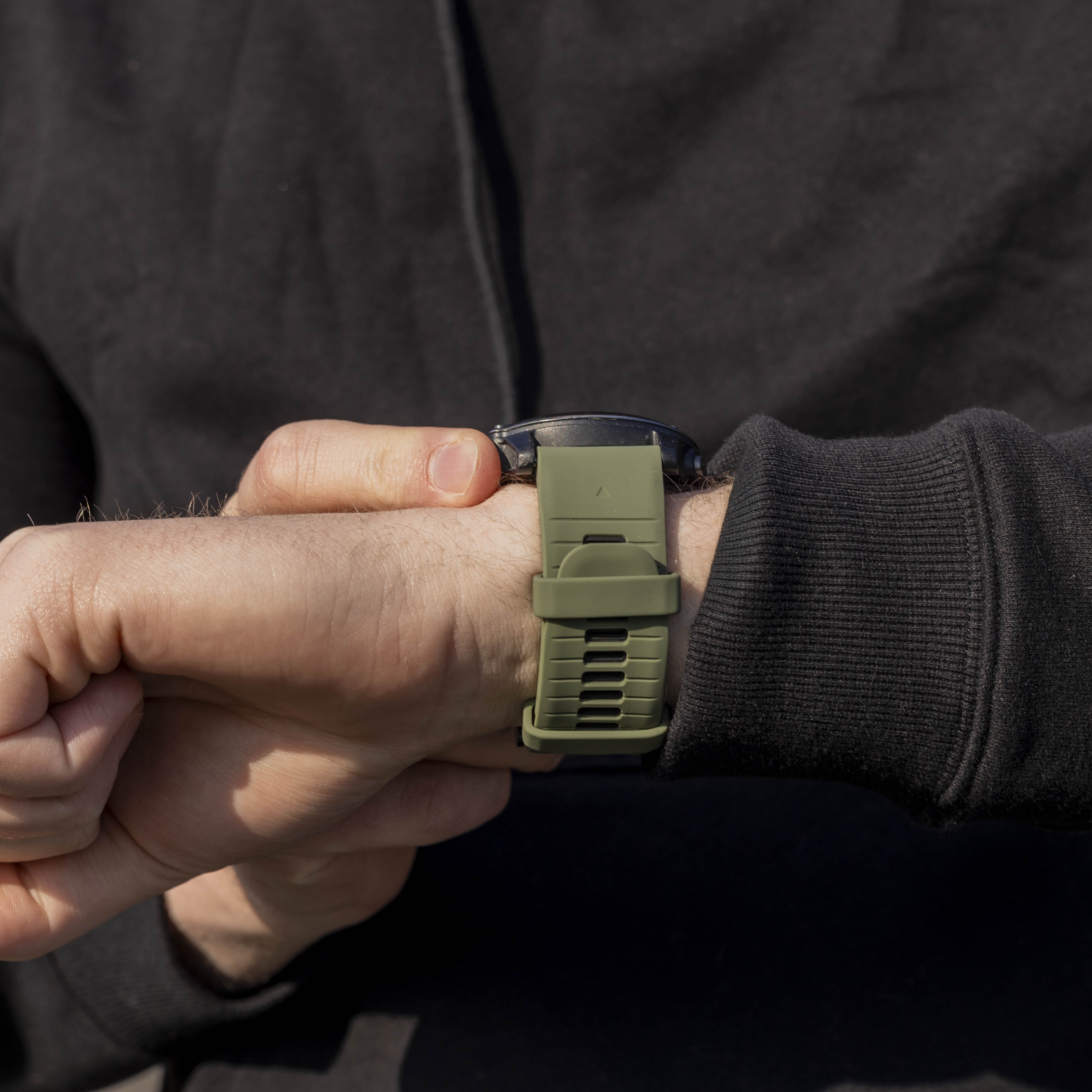 Nahaufnahme eines Handgelenks mit einer olivgrünen Uhr. Die Person trägt einen schwarzen Kapuzenpullover. Das Uhrenarmband ist befestigt.