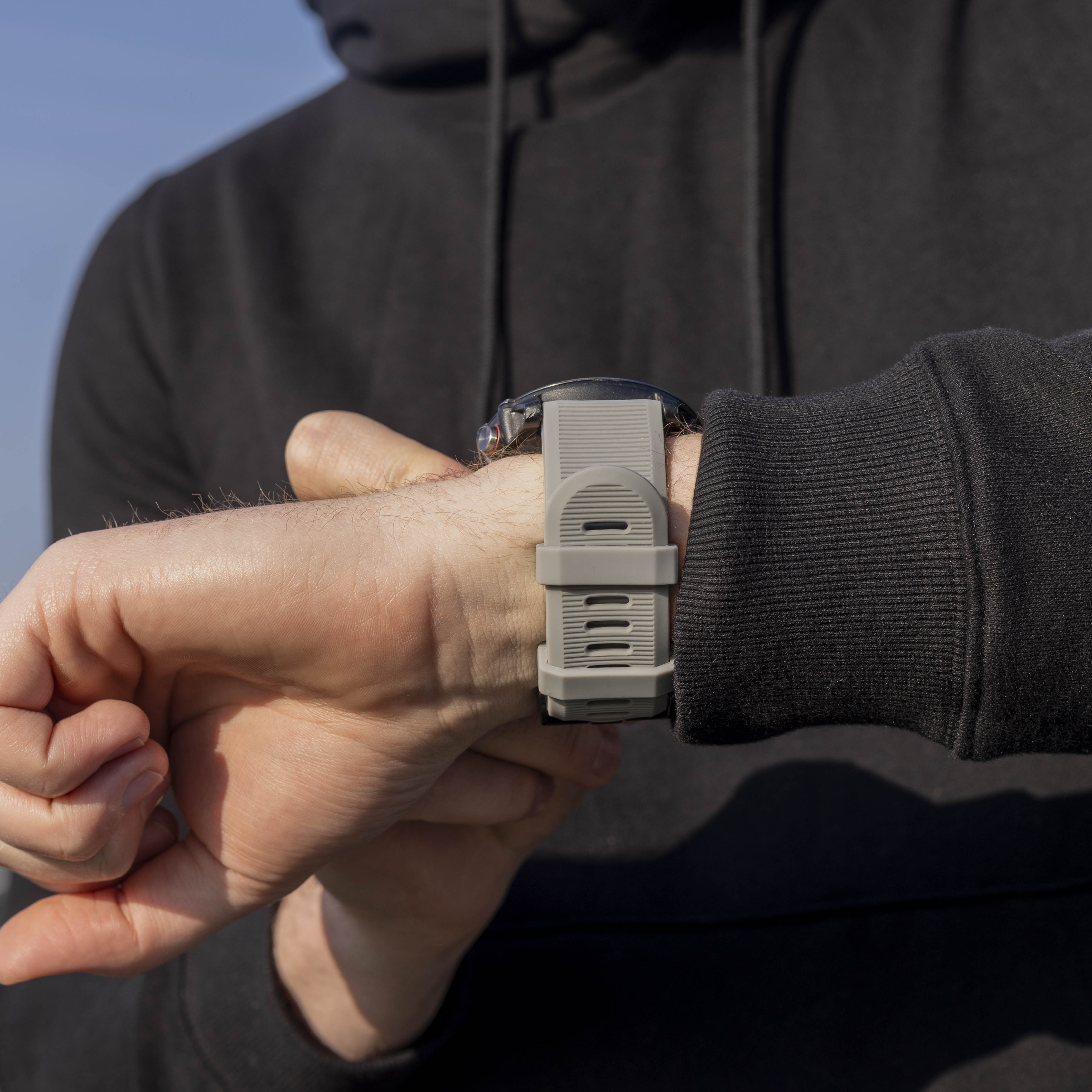 Person mit schwarzem Kapuzenpullover, die die Zeit auf einer grauen Smartwatch abliest. Blauer Himmel im Hintergrund sichtbar.