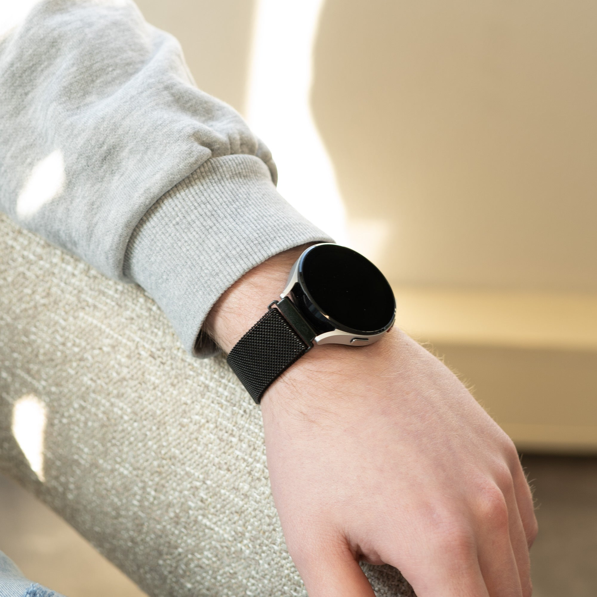 Handgelenk einer Person mit schwarzer Smartwatch. Die Uhr hat einen schwarzen Bildschirm und ein schwarzes Mesh-Armband. Die Person trägt einen grauen Pullover.