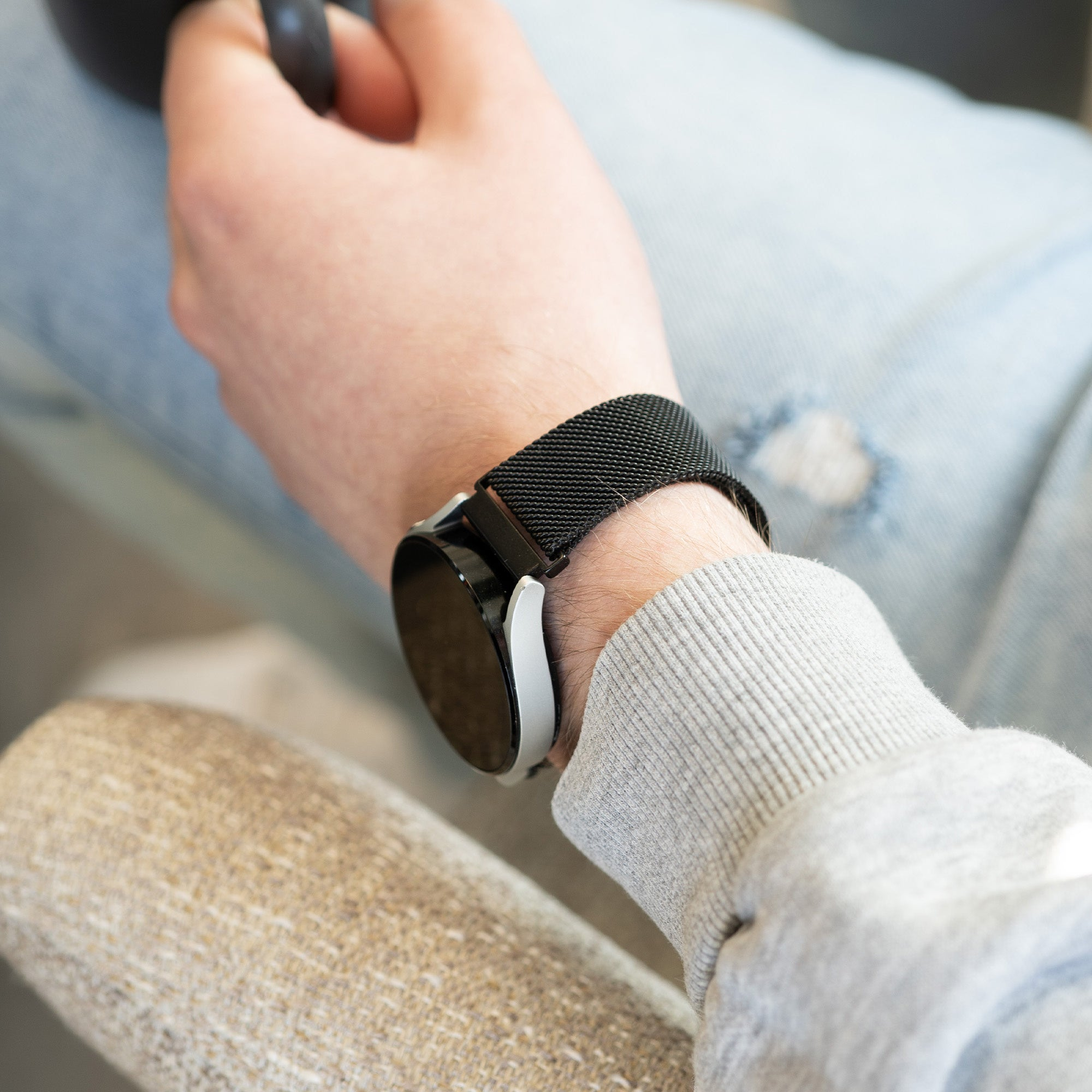 Das Handgelenk einer Person mit einer Smartwatch. Die Uhr hat ein schwarzes Armband, die Person trägt einen grauen Pullover.