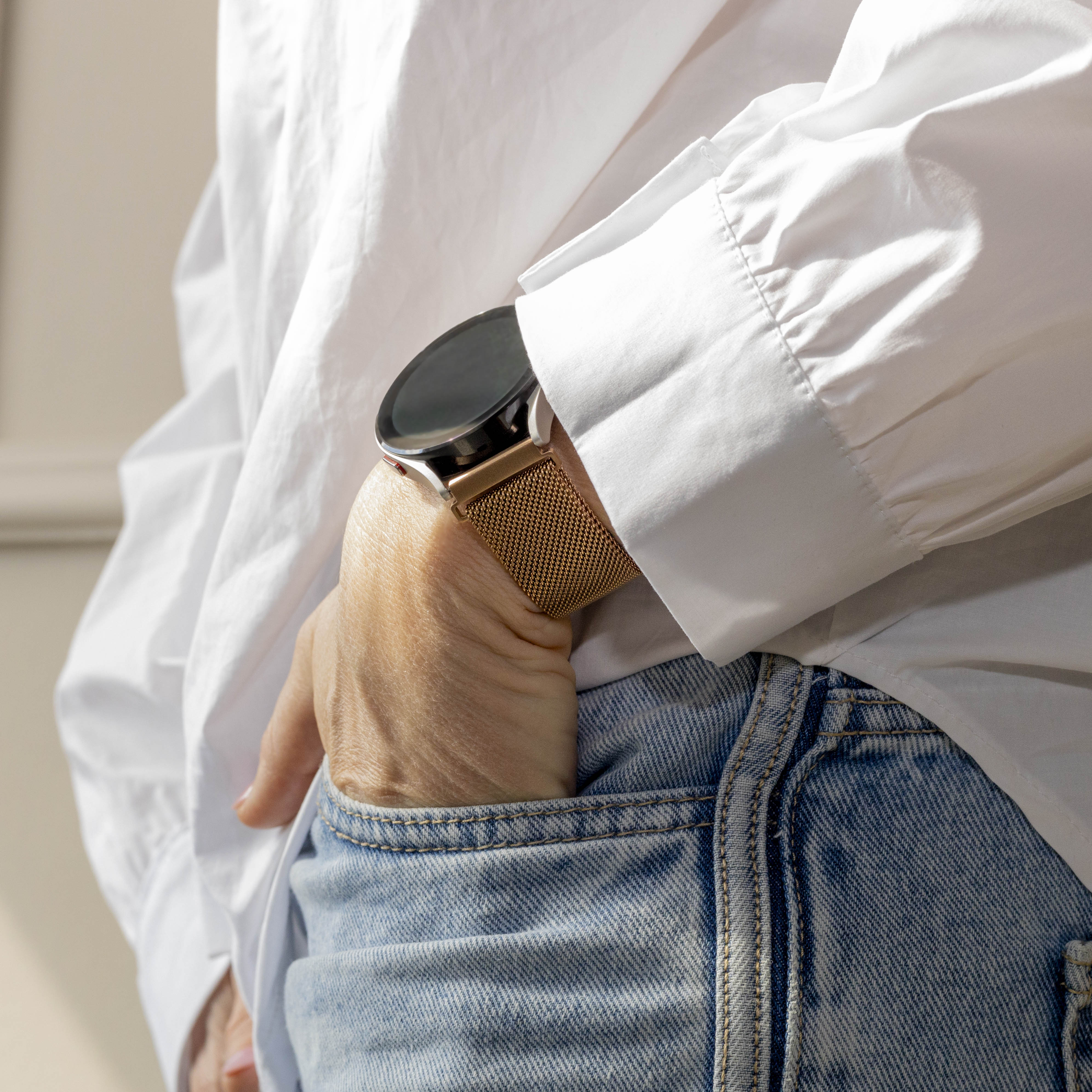 Eine Person trägt ein weißes Hemd und blaue Jeans. Am Handgelenk befindet sich eine goldene Uhr. Hand in der Tasche.