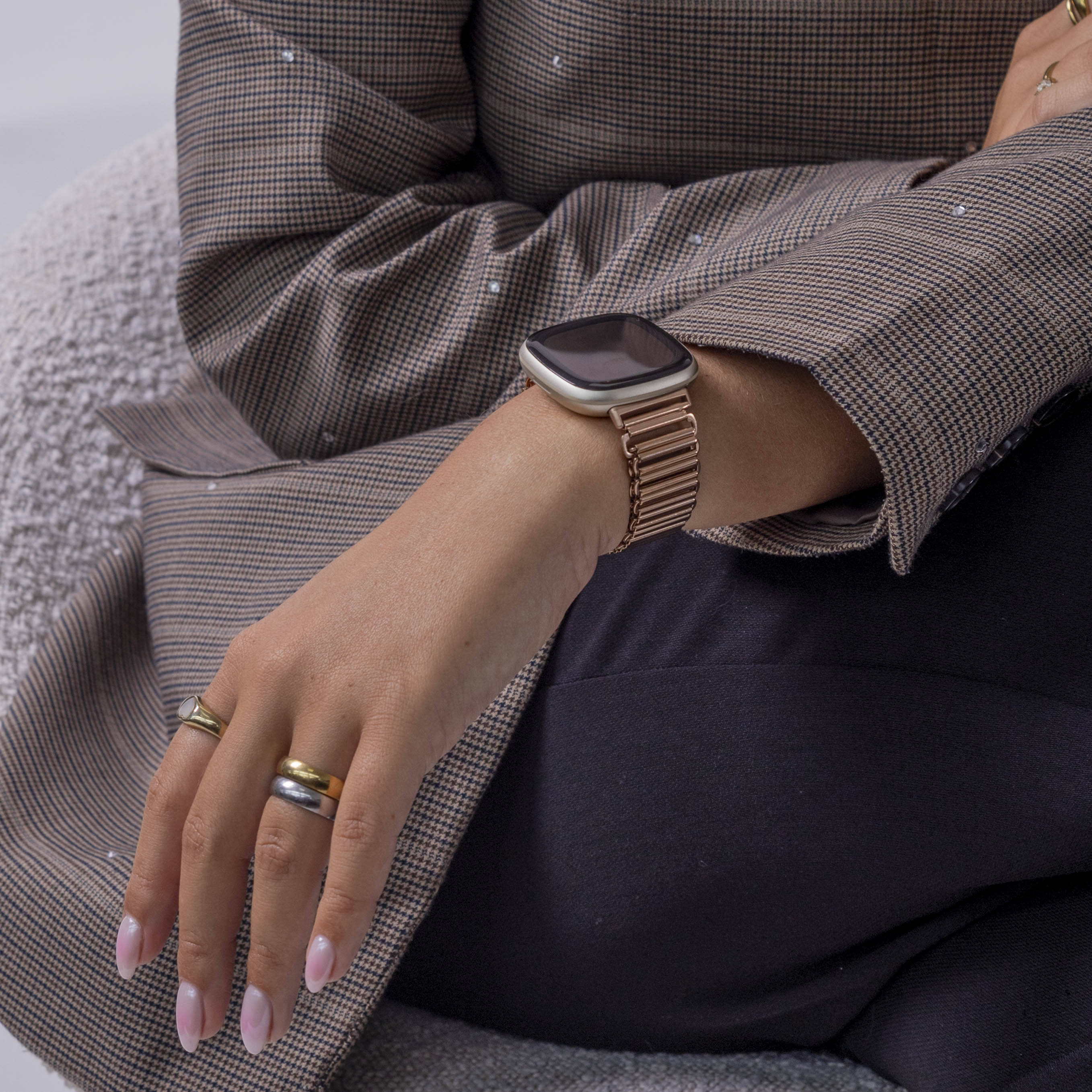 Person trägt Uhr am Handgelenk, Ringe und einen braunen Blazer mit Muster. Schwarze Hose ist sichtbar. Der Hintergrund ist unscharf.