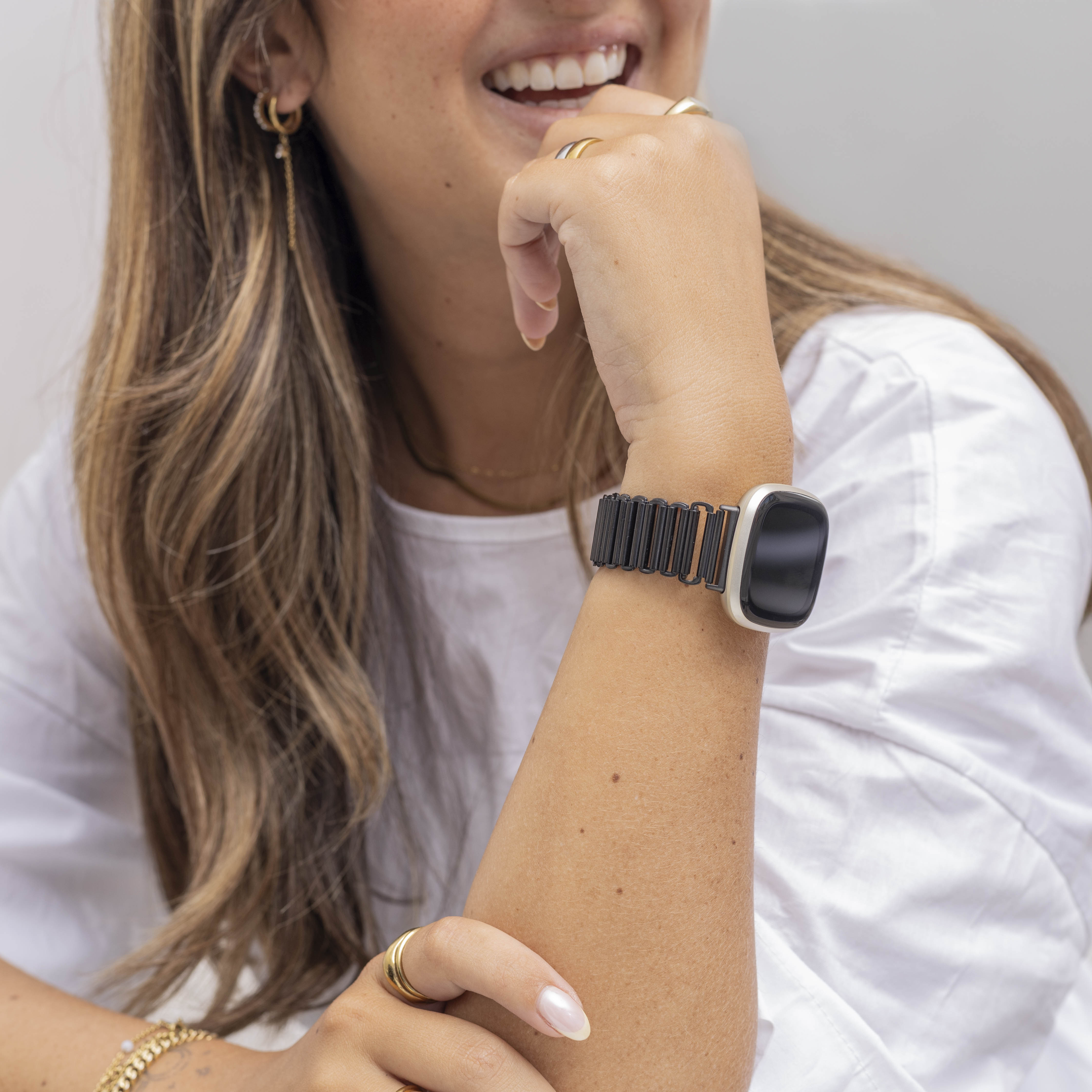 Eine lächelnde Frau mit langen Haaren trägt eine Armbanduhr. Sie trägt Goldschmuck und ein weißes Oberteil. Ihre Hand ist in der Nähe ihres Mundes.