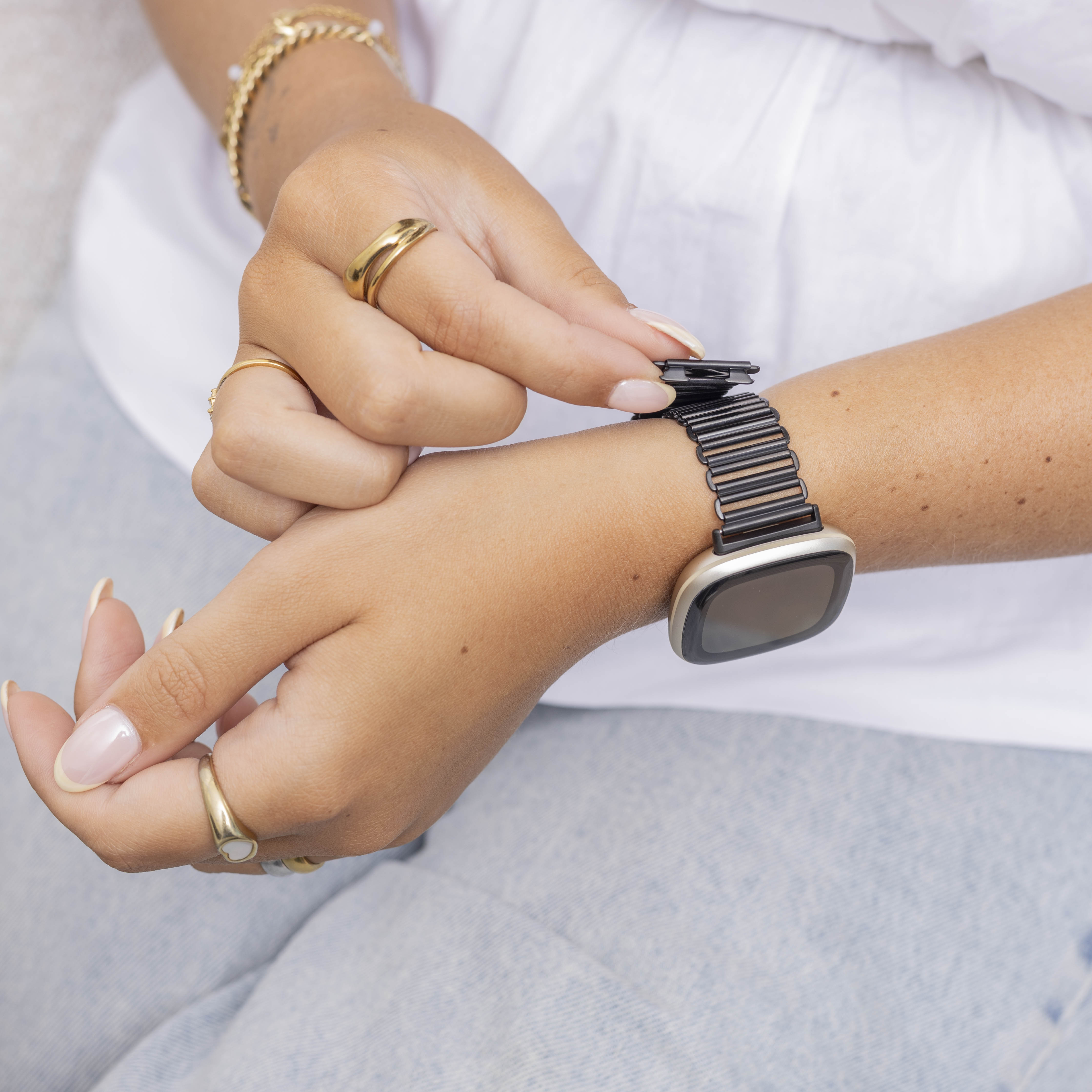 Eine Person stellt eine schwarze Uhr an ihrem Handgelenk ein. Sie trägt Ringe, hat Manikürenägel und ein weißes Hemd.