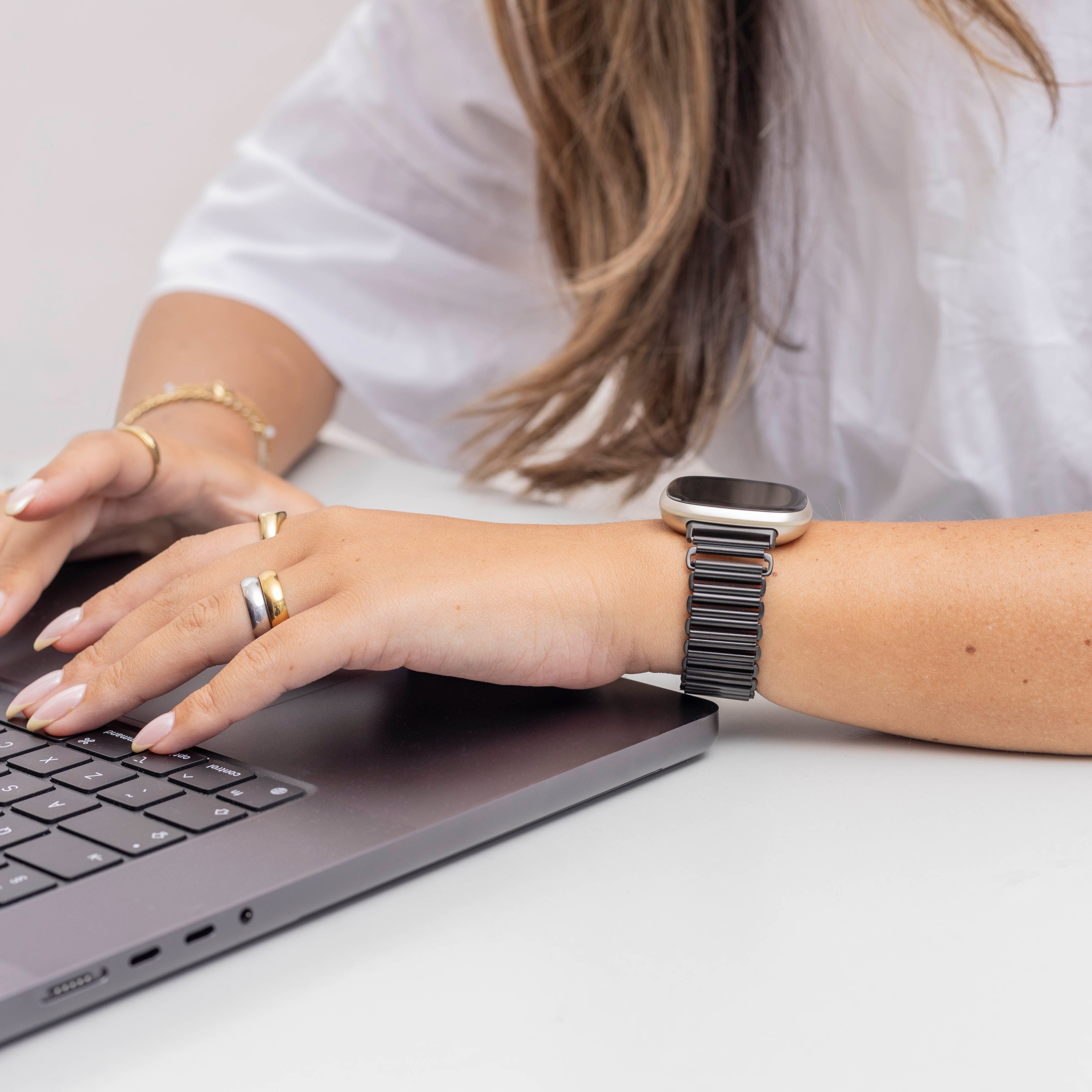 Eine Person tippt auf einem Laptop. Sie trägt Ringe und eine Uhr mit schwarzem Armband. Weißes Hemd und heller Hintergrund.