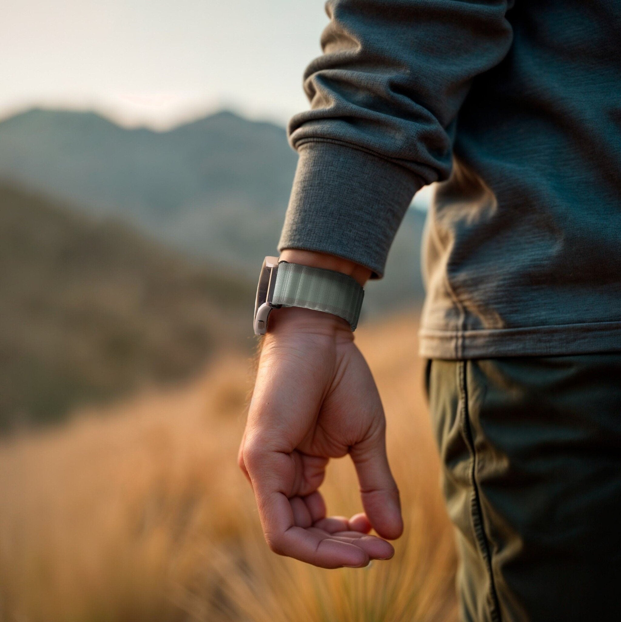 Arm und Hand einer Person mit Smartwatch. Im Hintergrund sind Berge und ein Feld mit hohem Gras zu sehen.