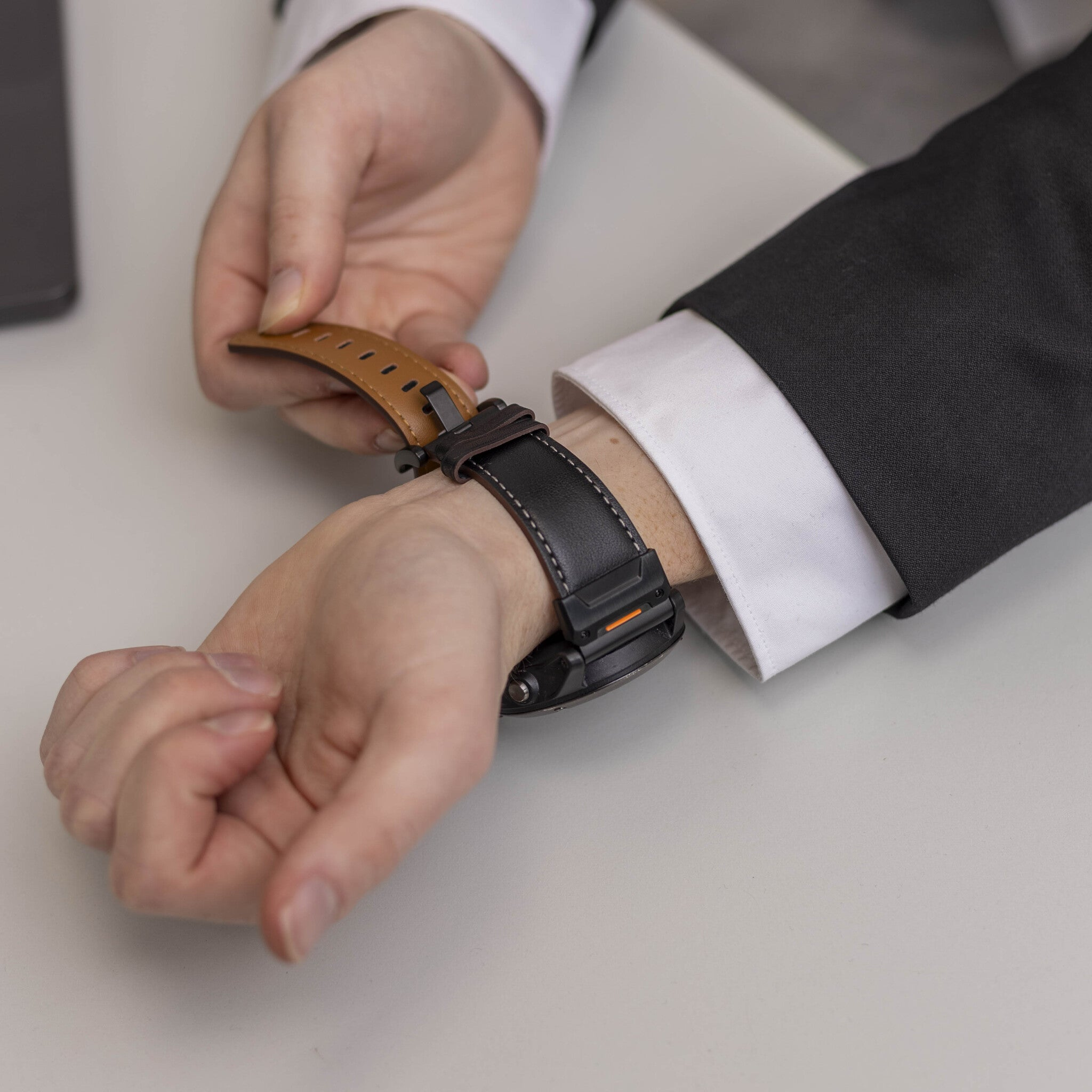 Eine Person im Anzug befestigt eine schwarze Uhr mit einem braunen Lederarmband am Handgelenk.