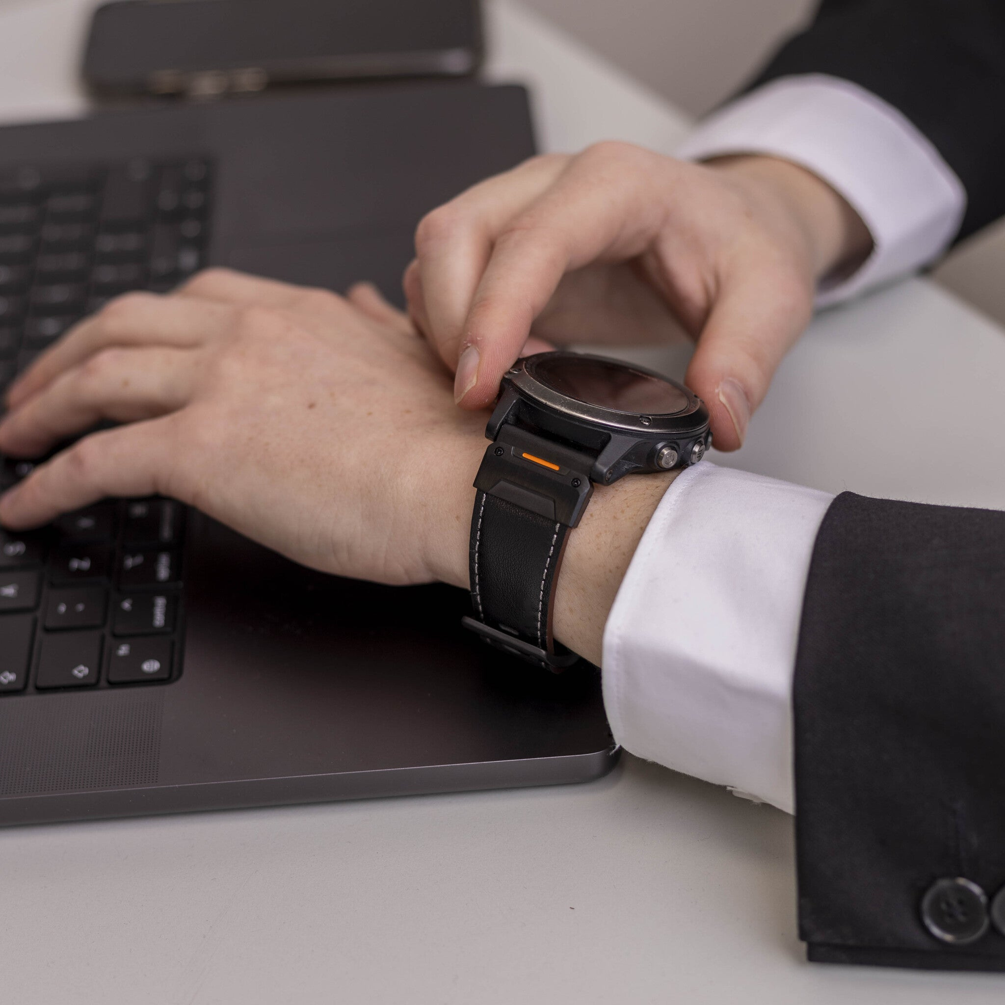 Eine Person im Anzug justiert eine Smartwatch. Die Uhr hat ein schwarzes Lederarmband und befindet sich am Handgelenk. Ein Laptop ist sichtbar.
